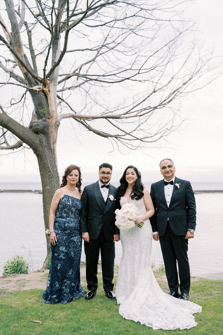 Four elegantly dressed people pose by a tree on a lakeside setting, with the bride in a stunning white lace gown holding a bouquet.