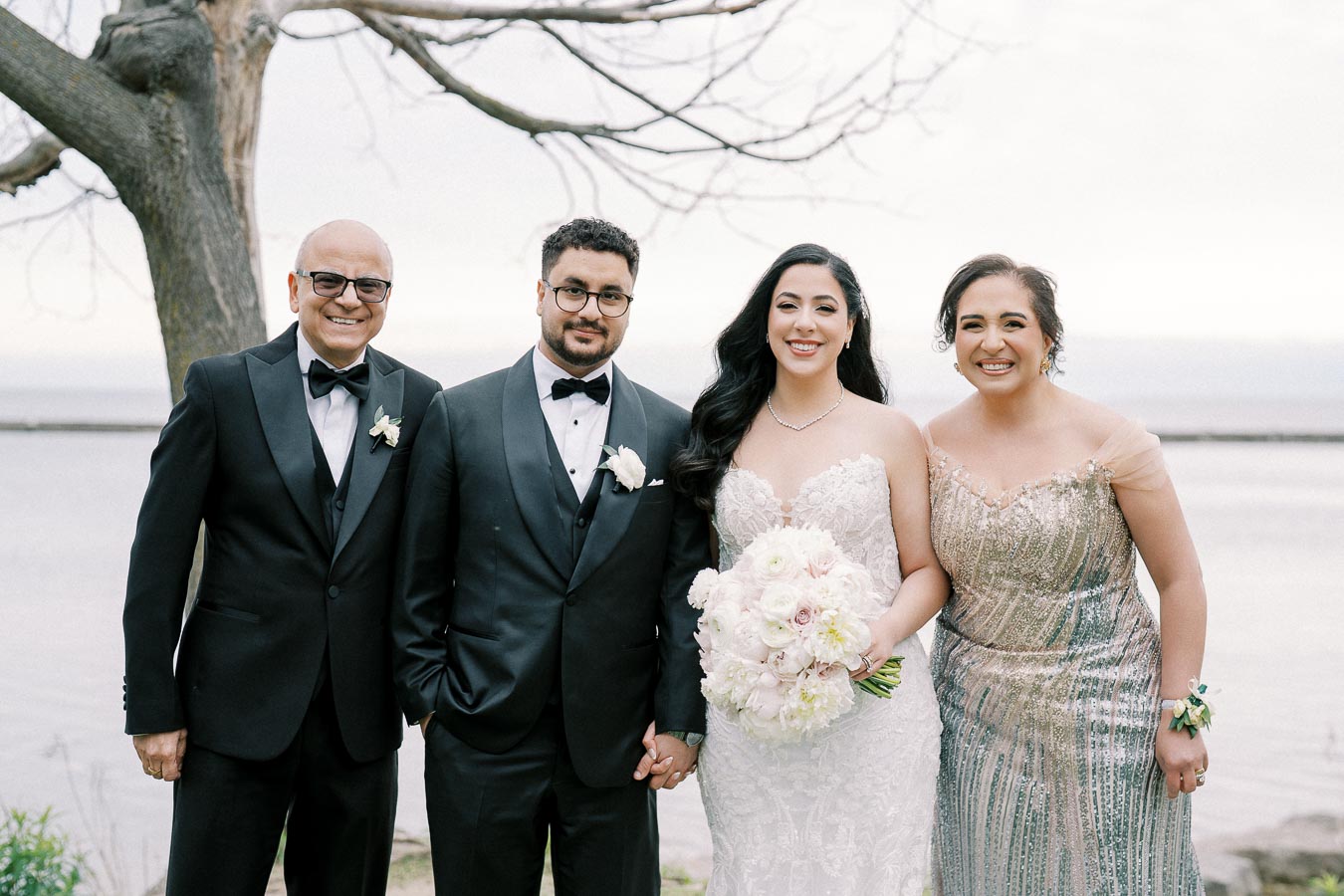 A happy bride and groom pose with their parents by a scenic lakeside, celebrating a beautiful outdoor wedding ceremony.