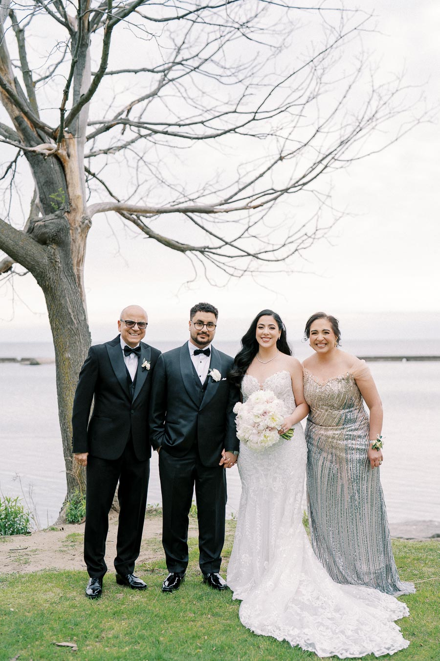Wedding couple posing with parents by a lake, dressed elegantly; bride in a lace gown holding a bouquet, groom and father in tuxedos, mother in a sequined dress, tree and calm water in the background.