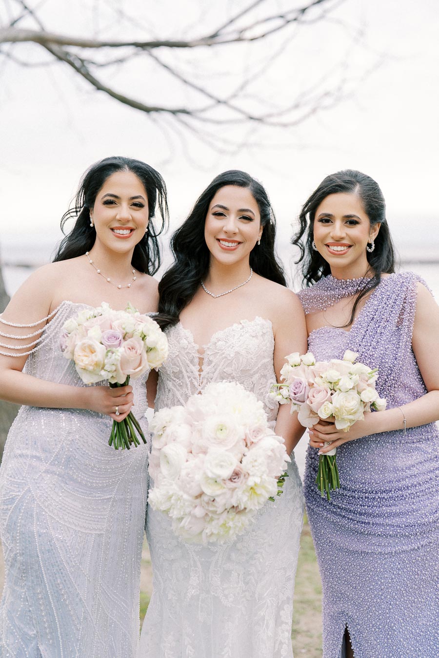 Three women in elegant gowns pose outdoors, holding pastel bouquets. The central woman wears a white lace wedding dress, flanked by bridesmaids in lavender and lilac sequin gowns, smiling under a cloudy sky with bare tree branches in the background.