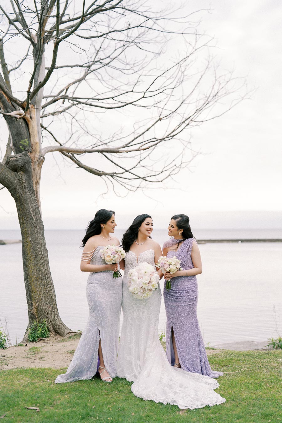 Three women in elegant dresses holding bouquets, standing by a tree next to a serene lake.