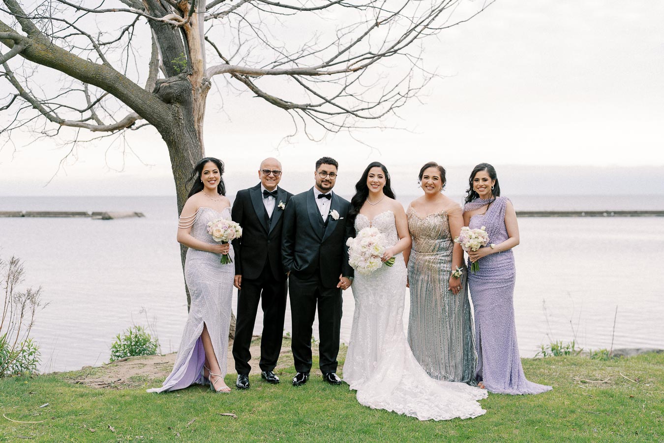 Wedding party posing by a lakeside under a leafless tree, featuring a bride in a white gown, groom in a black tuxedo, and wedding guests in elegant dresses, all holding floral bouquets.