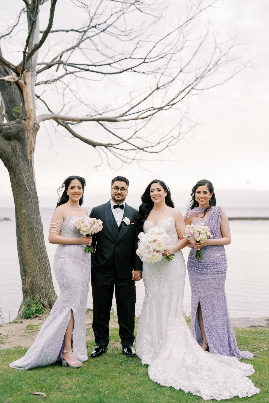 Wedding party posing by a lakeside, featuring the groom in a black tuxedo and three bridesmaids in elegant lilac and white gowns holding floral bouquets, with a tree and calm water in the background.