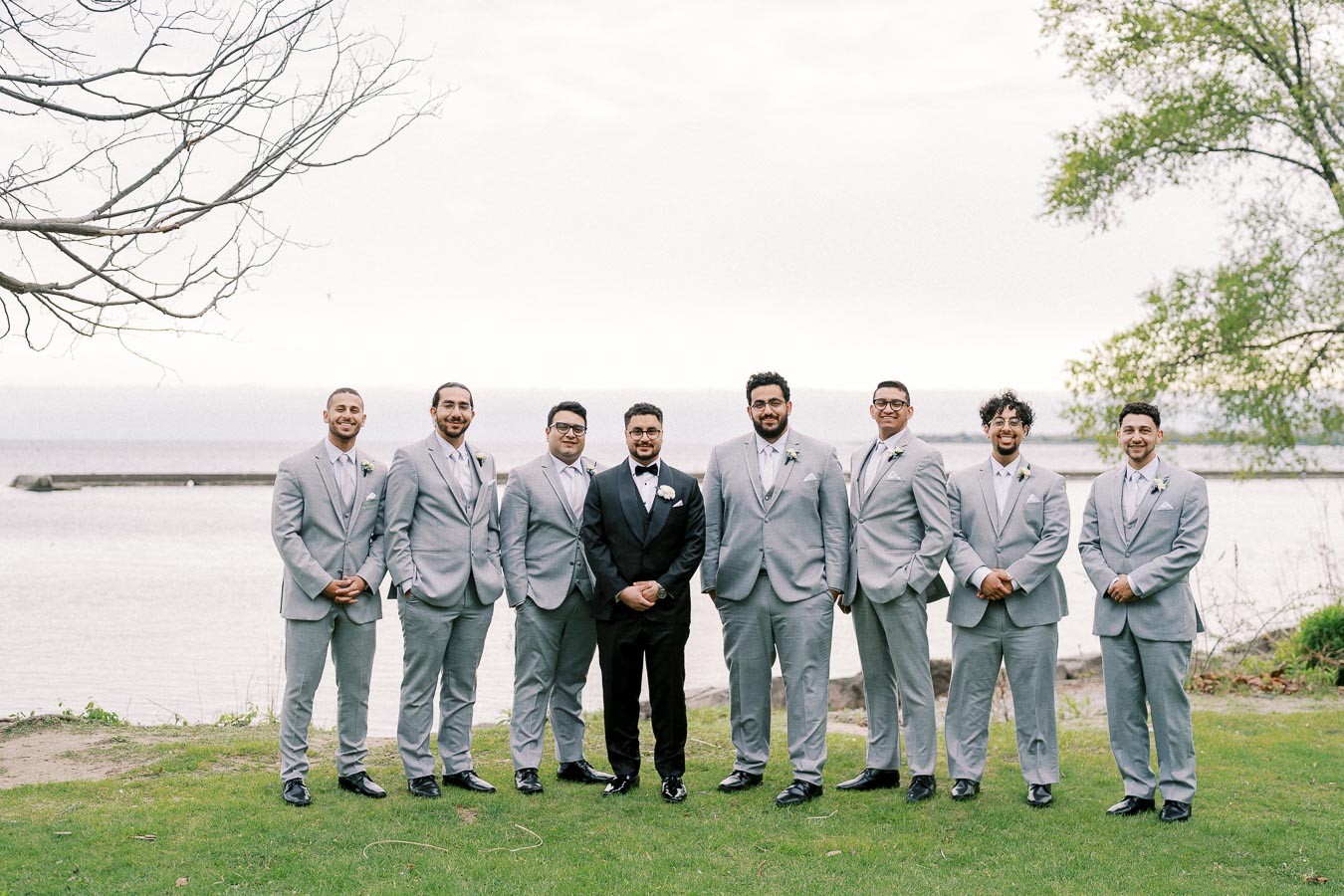A group of eight groomsmen in light gray suits stand on a grassy area by a lake, with a groom in a black tuxedo in the center, under a cloudy sky with trees framing the scene.
