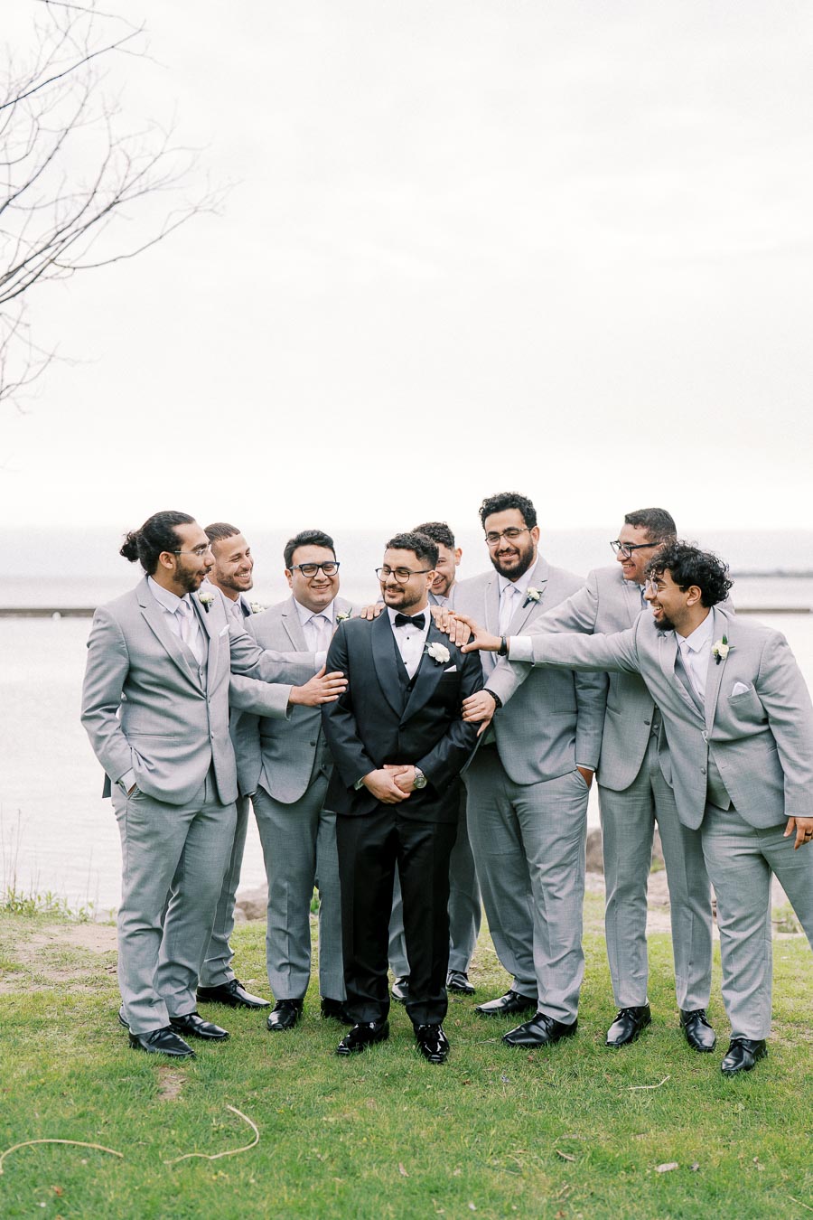 Groom in a black tuxedo standing with groomsmen in matching gray suits, smiling and celebrating outdoors with a lake in the background.