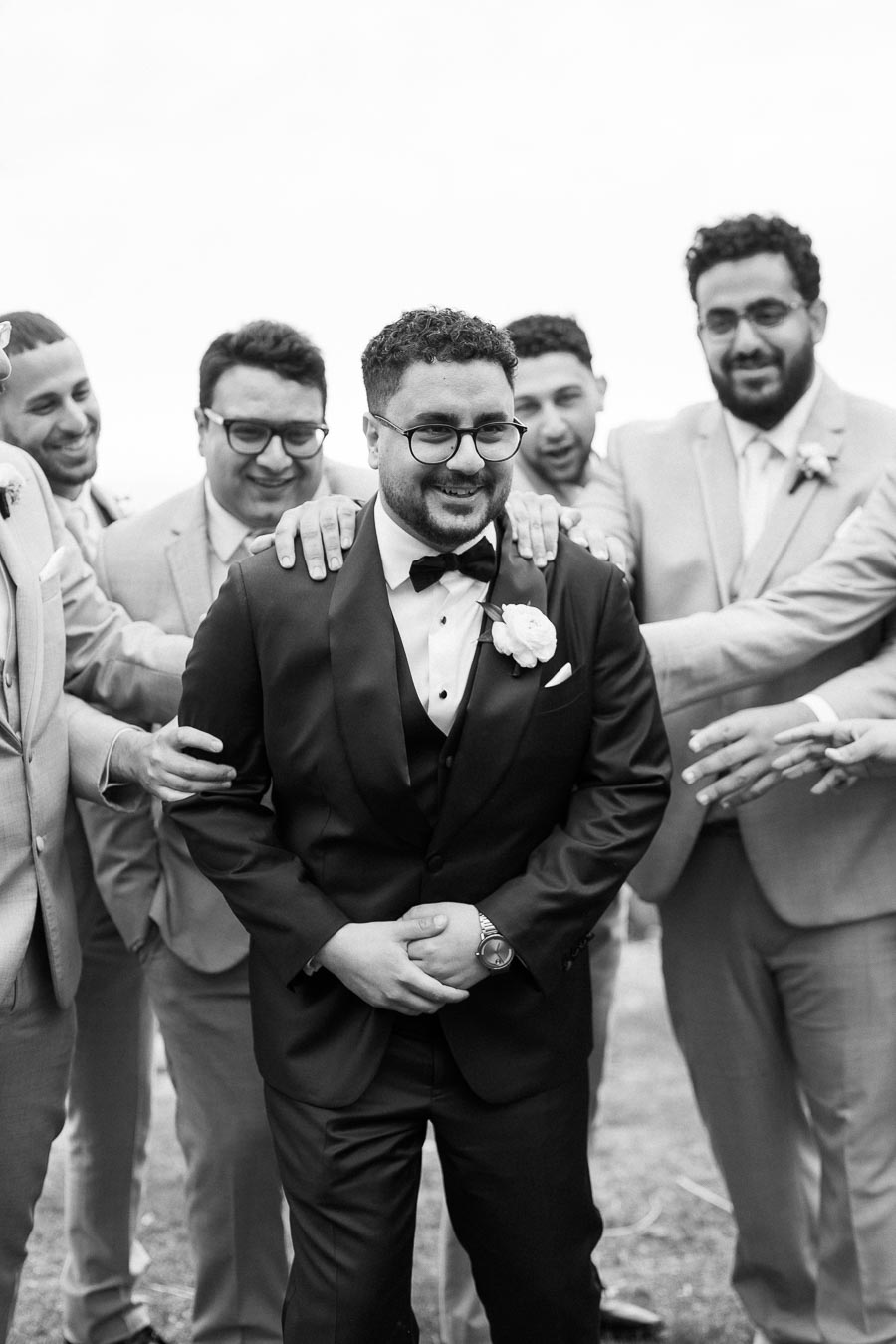 A group of groomsmen in suits sharing a joyful moment with the groom, all smiling and standing together. The groom is dressed in a black tuxedo with a bow tie and a white boutonniere, while his friends support him with hands on his shoulders. Black and white photo captures the camaraderie and happiness at a wedding event.