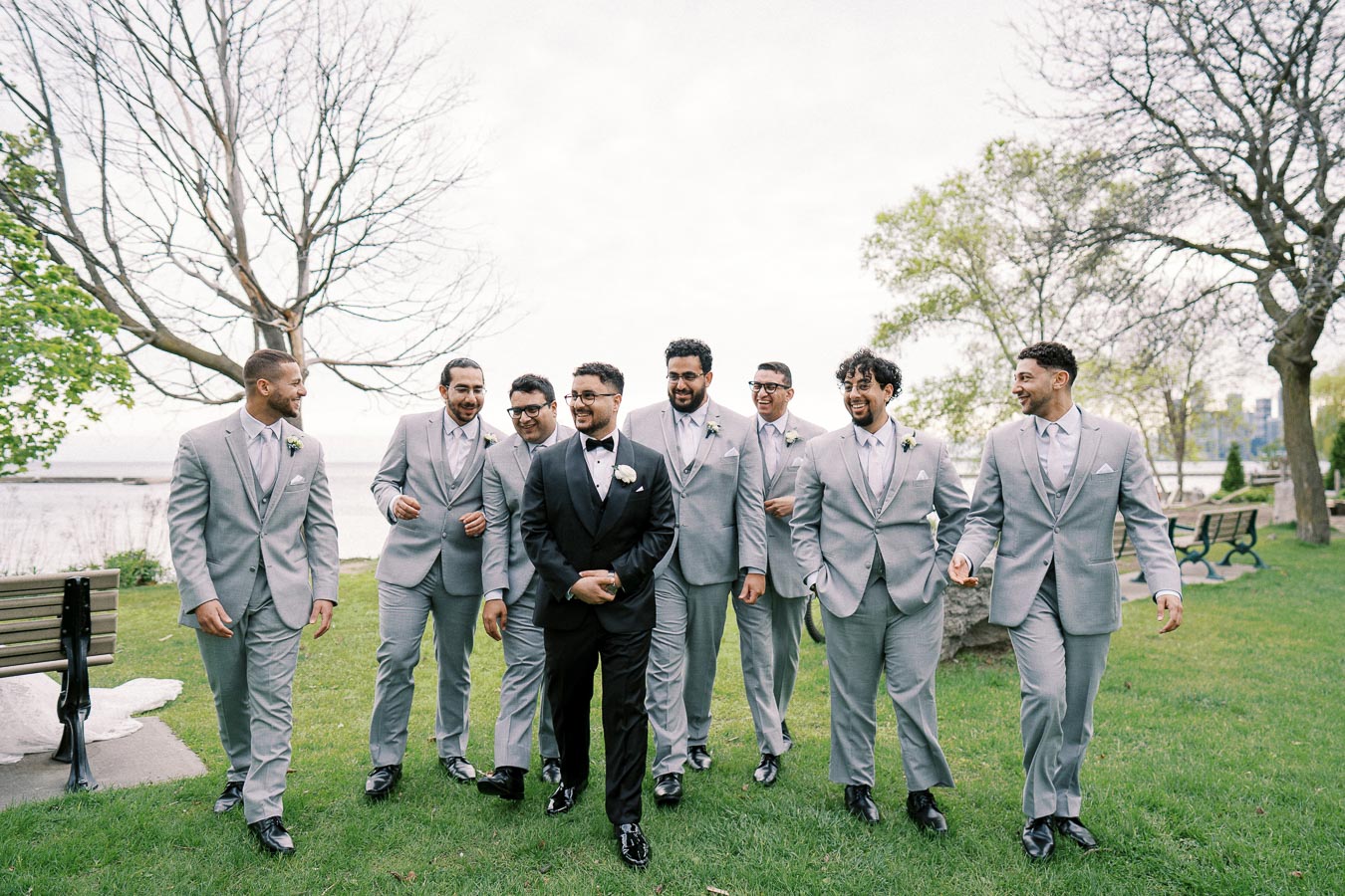 A groom in a black tuxedo walks with his groomsmen in light gray suits in a park by the water, surrounded by trees and greenery on a wedding day.