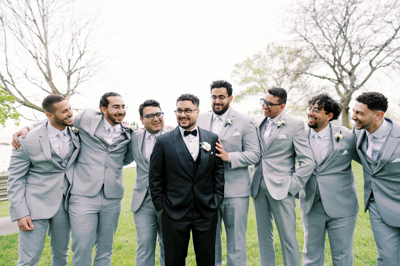 Groom in a black tuxedo smiles while standing with seven groomsmen in light gray suits, posing jovially outdoors on a lush green lawn with trees in the background.