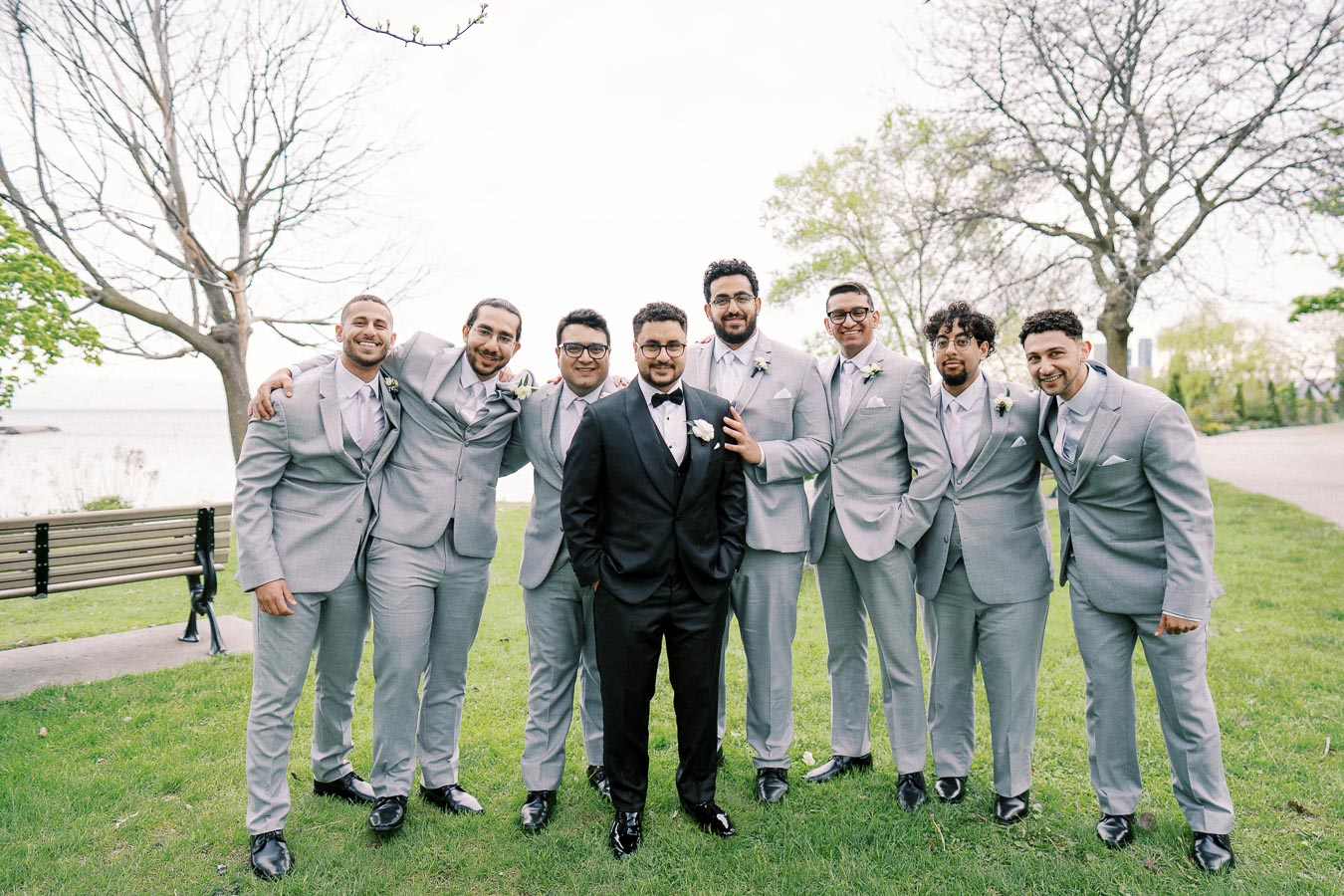 A group of groomsmen in light gray suits and a groom in a black tuxedo pose together in a park setting with trees and a bench, showcasing a wedding celebration atmosphere.