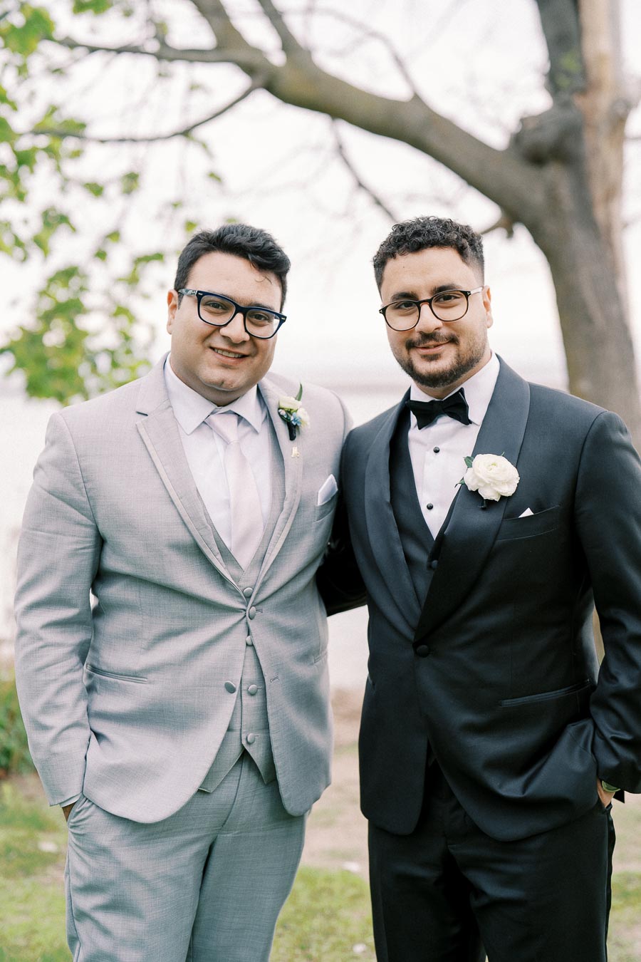 Two men in formal attire, one in a light gray suit and the other in a black tuxedo, smiling and posing together outdoors in a park-like setting.