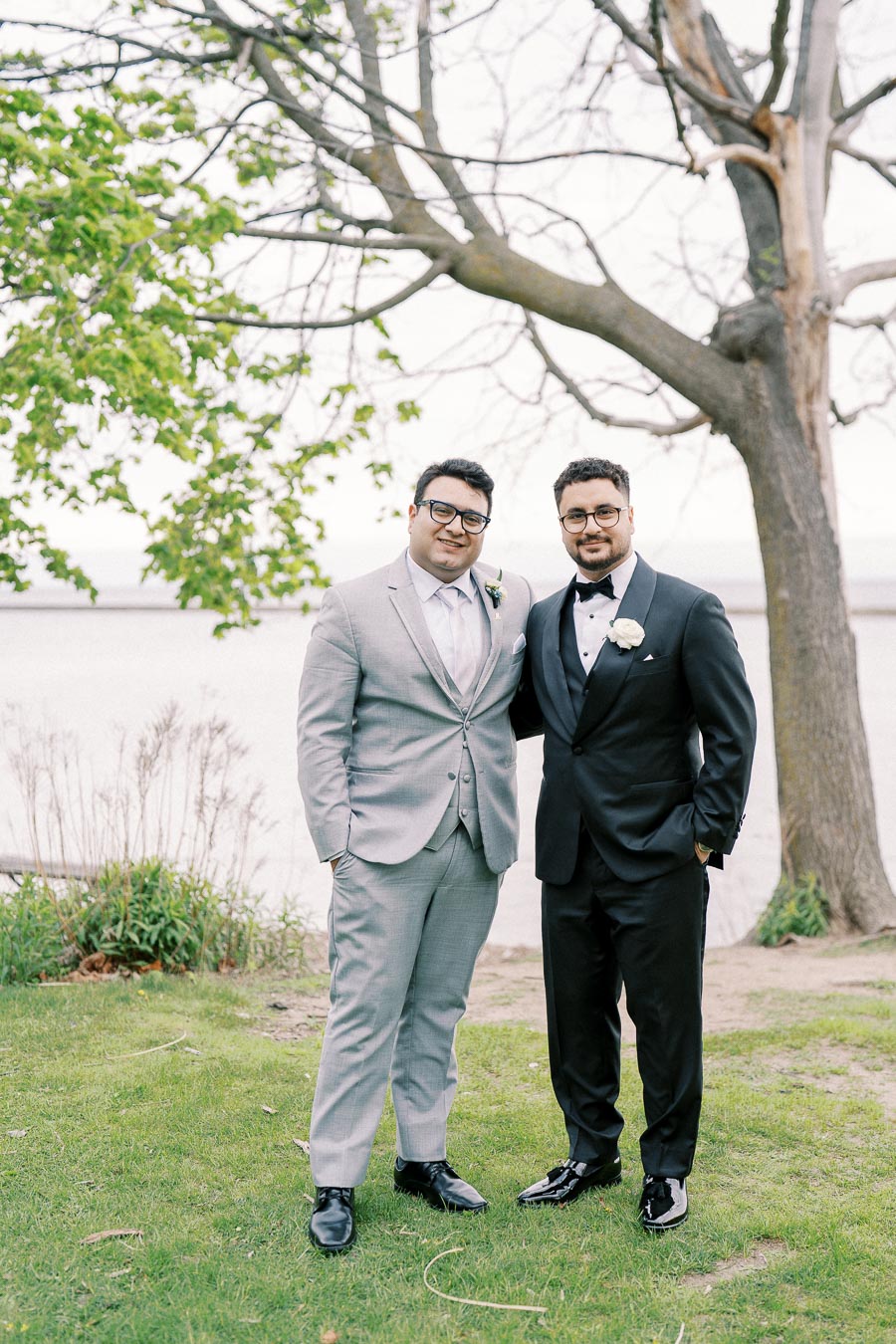 Two men wearing formal attire, one in a light gray suit and the other in a black tuxedo, pose together outdoors near a tree and a body of water. They are standing on green grass, with a backdrop of natural scenery, including a tree with budding leaves.
