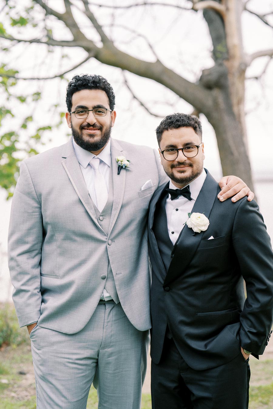 Two men in formal suits standing together outdoors, one in a light gray suit and the other in a black tuxedo, both wearing boutonnieres, with a tree in the background.