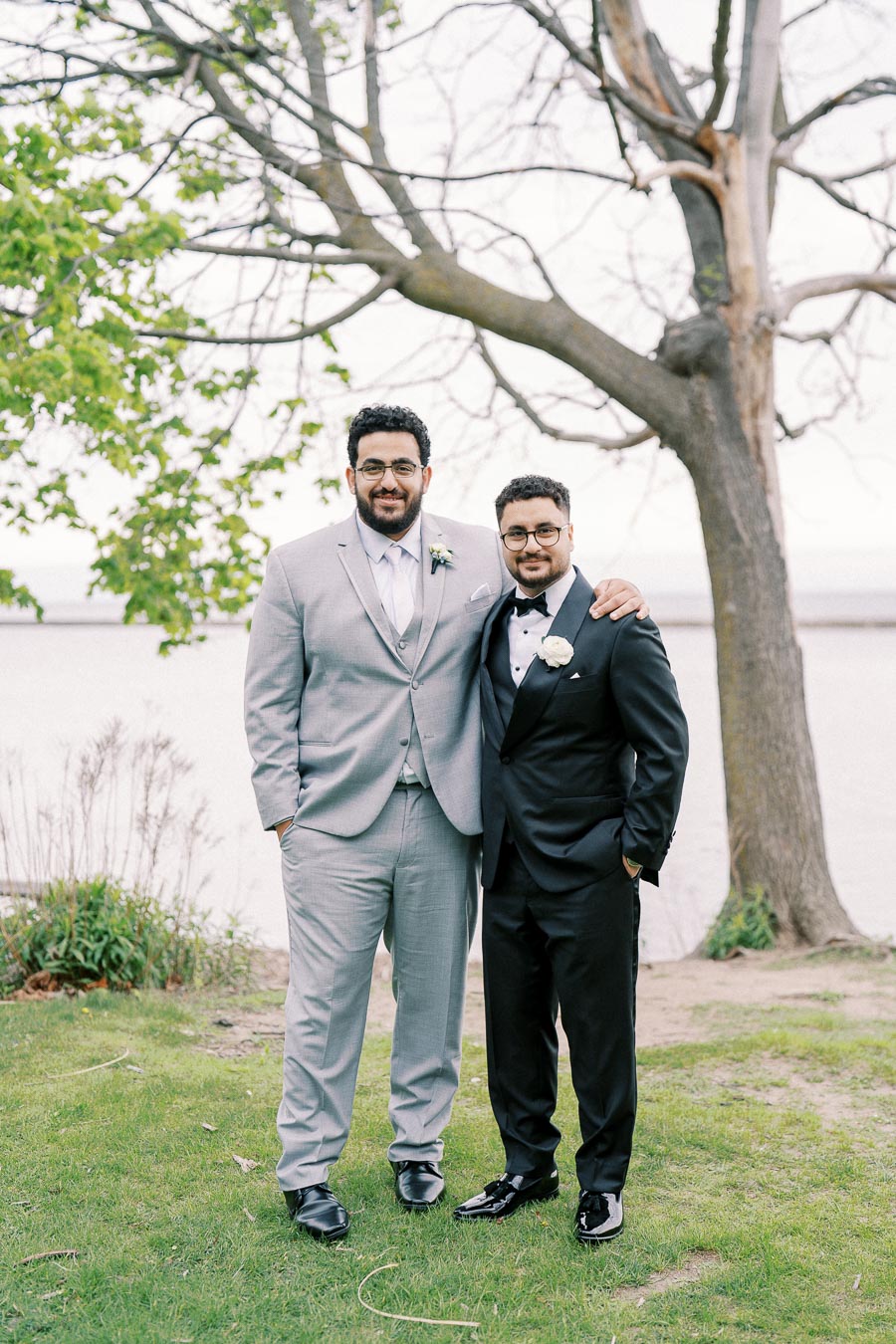 Two men in formal attire, one in a gray suit and the other in a black tuxedo, standing on grass near a leafless tree by a lake.