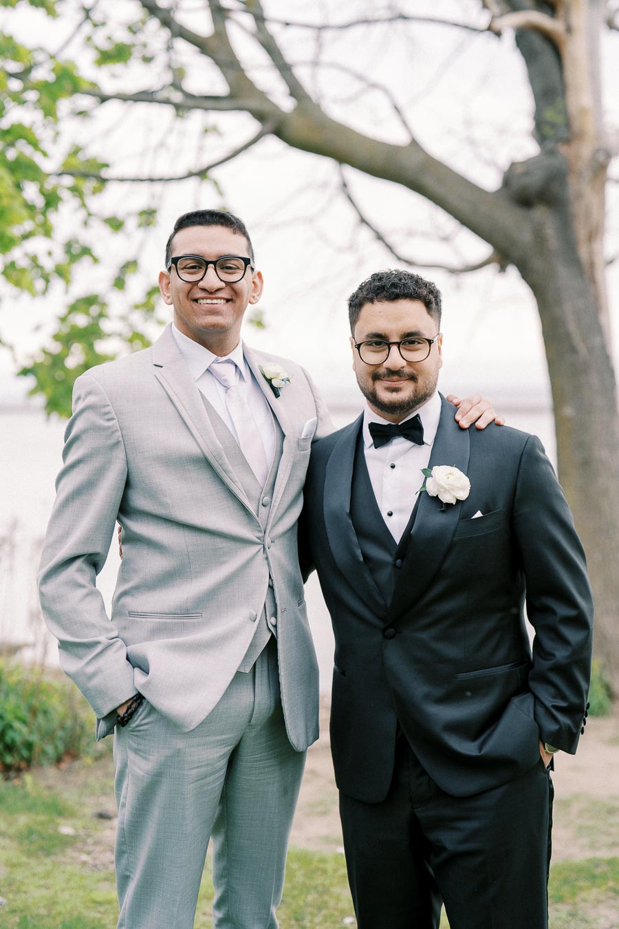 Two men wearing formal suits, one in grey and the other in black, smiling and posing together in an outdoor setting with trees and greenery in the background.