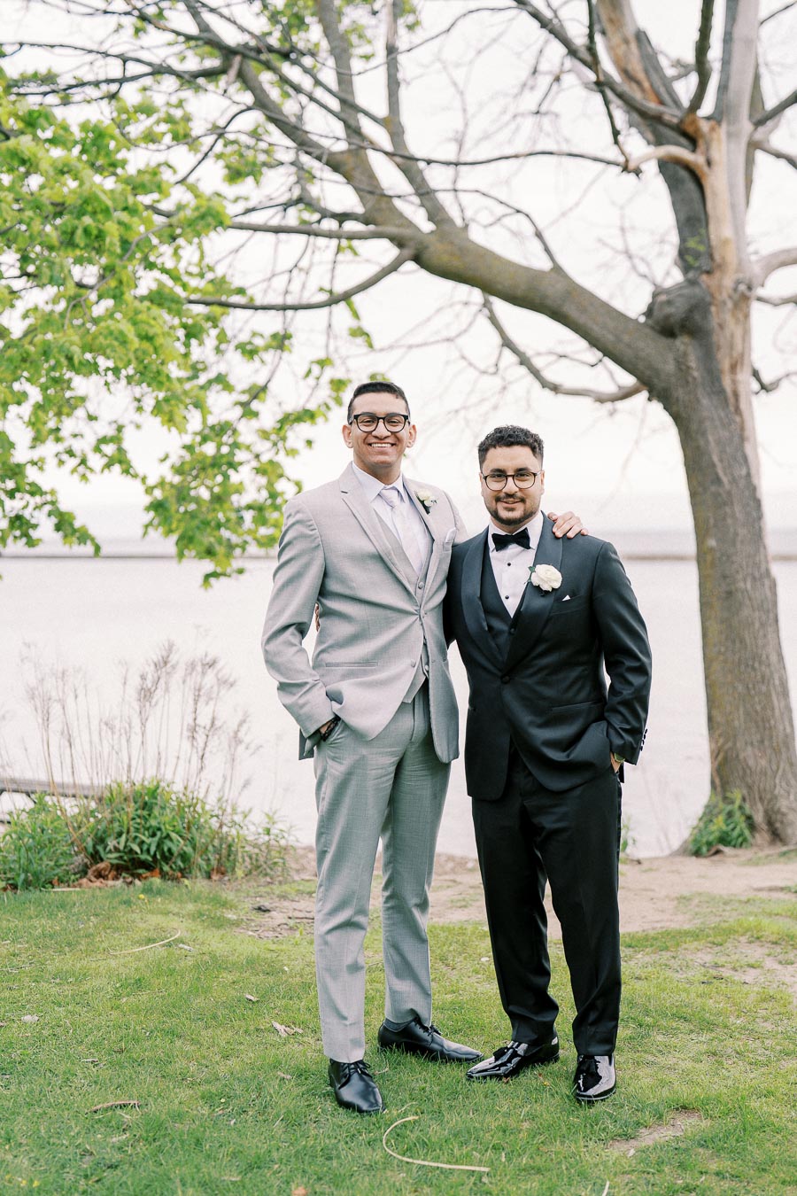 Two men in formal suits posing outdoors by a lake and tree, with a natural green and serene backdrop, capturing a moment of friendship and celebration at a wedding or formal event.