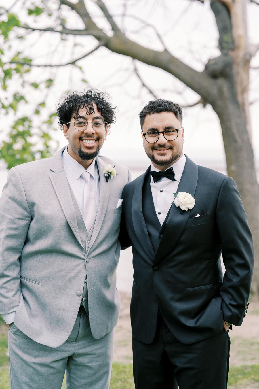 Two men wearing suits, one in gray and one in black, smiling at a wedding ceremony outdoors with a tree and greenery in the background.