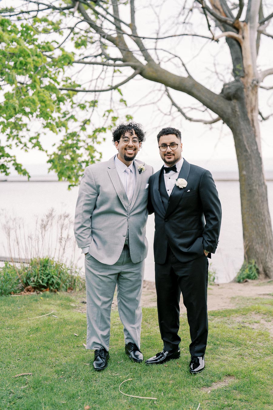 Two men dressed in formal suits, one gray and one black, standing outdoors on a grassy area by a large tree, smiling and posing for a photograph.