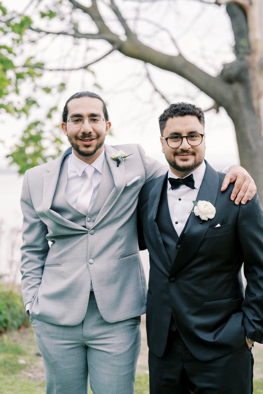 Two men in formal attire, one wearing a light gray suit and the other in a black tuxedo with a bow tie, pose together at an outdoor event. They each sport boutonnieres, suggesting a wedding or formal celebration, with a blurred natural background featuring trees.