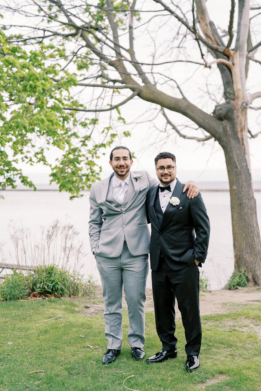 Two men in formal suits, one in light gray and the other in black, smiling and standing on grass with a tree and water in the background.