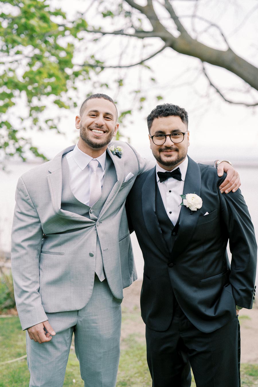 Groom and best man smiling in stylish suits outdoors at a wedding, with greenery in the background.