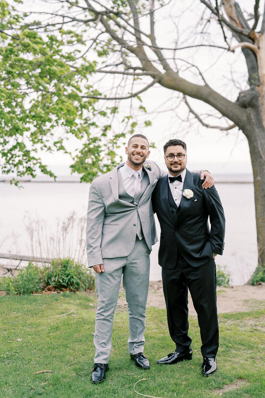 Two men dressed in formal suits standing outdoors on lush green grass by a lake, with one man in a light gray suit and the other in a black tuxedo, smiling under a tree.