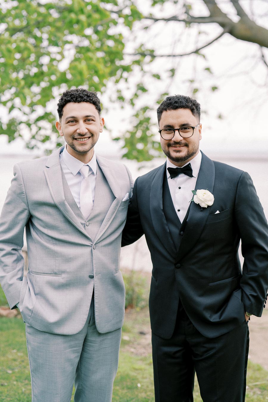 Two men smiling, one in a light gray suit and the other in a black tuxedo with a white boutonniere, standing outdoors with greenery in the background.