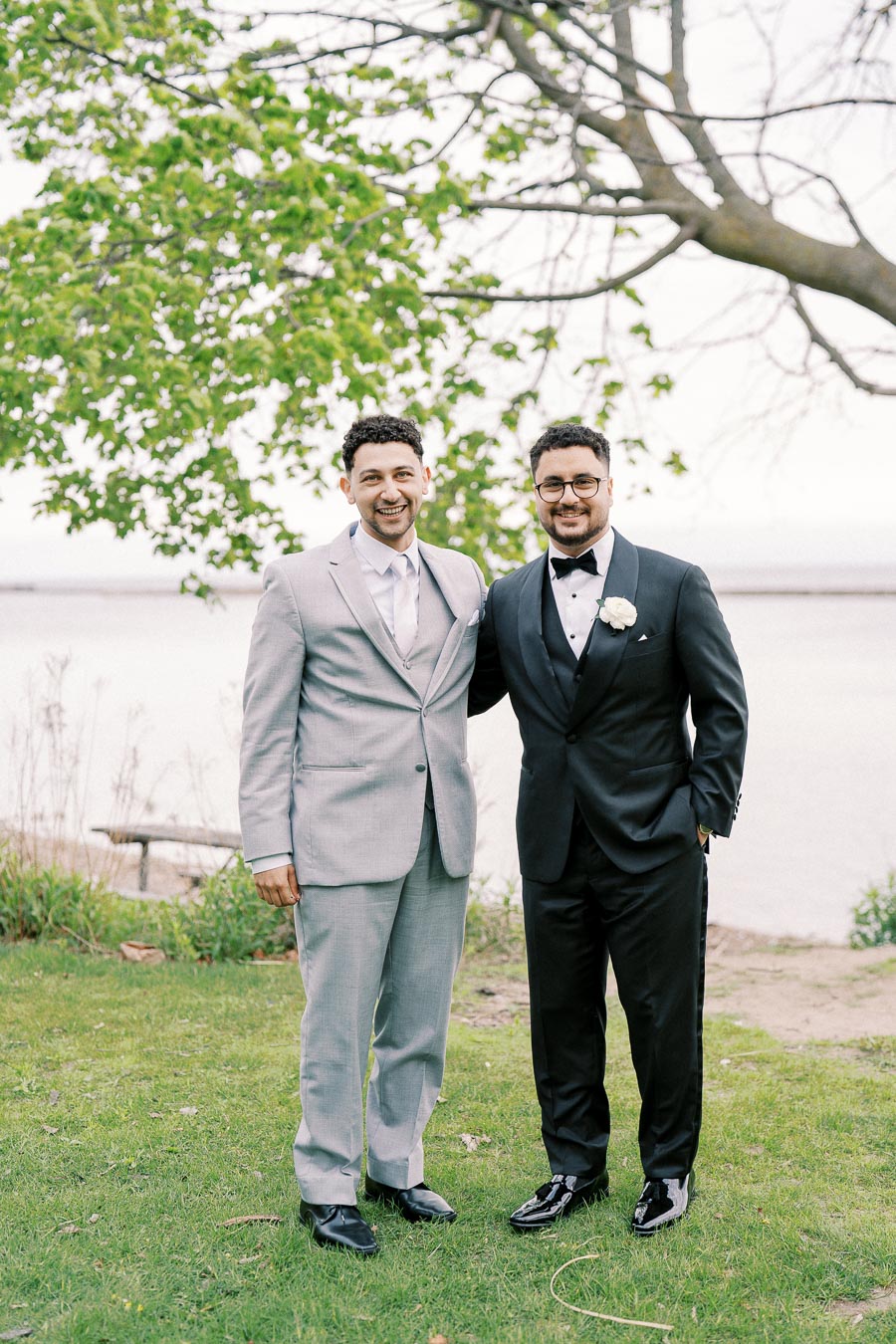 Two men in formal attire, one wearing a grey suit and the other a black tuxedo, standing together on a grassy area near a tree and a body of water, with smiles on their faces.