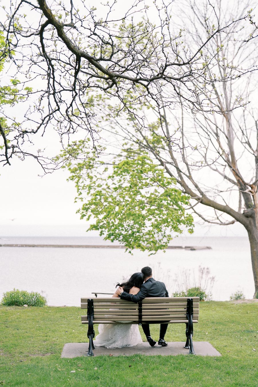 Couple in wedding attire sitting on a park bench by a lake, surrounded by lush spring trees and serene water.