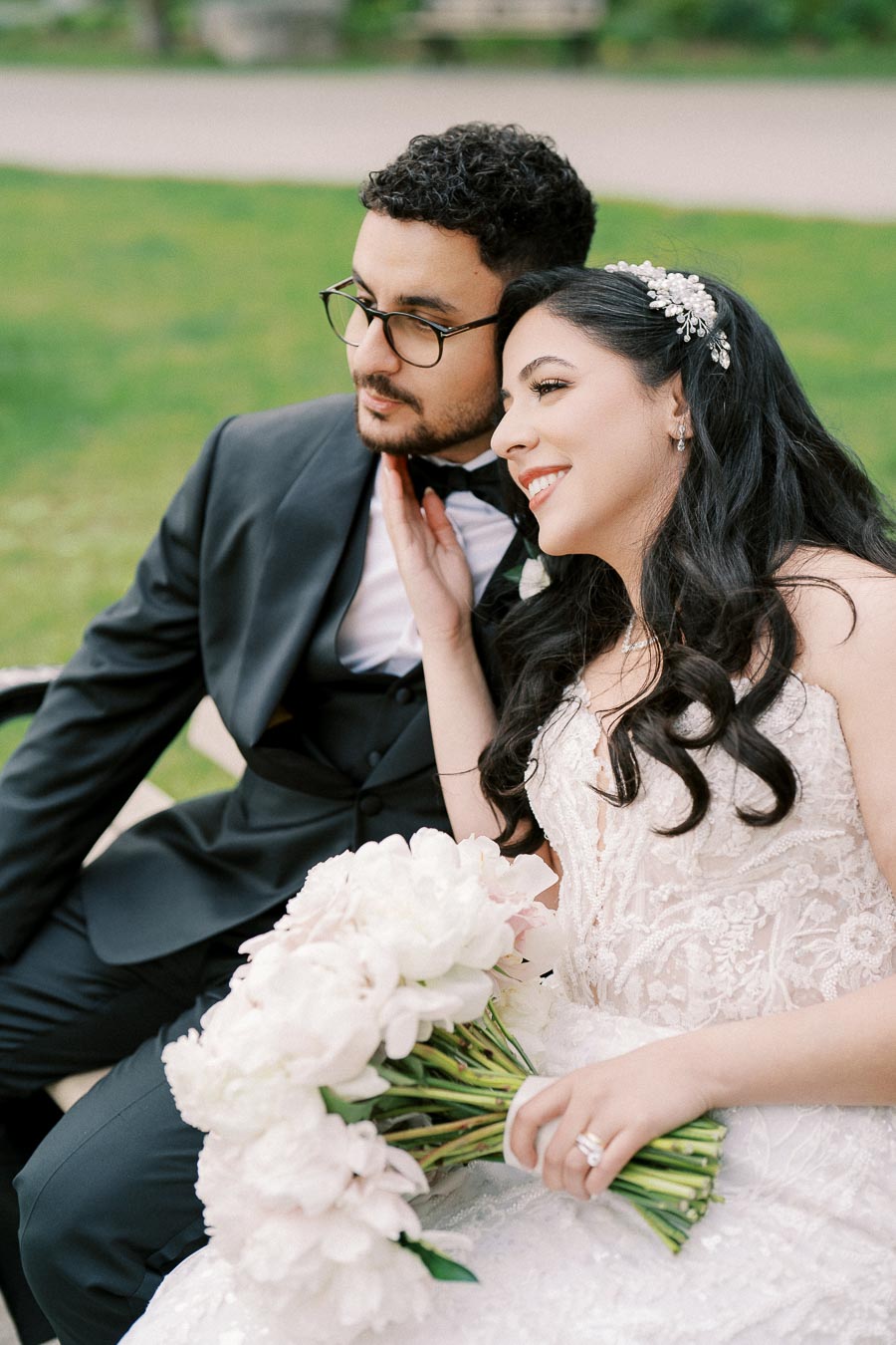 A bride and groom sitting together on a bench in a garden, the bride in a white lace wedding dress holding a bouquet of white flowers, and the groom in a black suit and glasses, sharing a joyful moment.