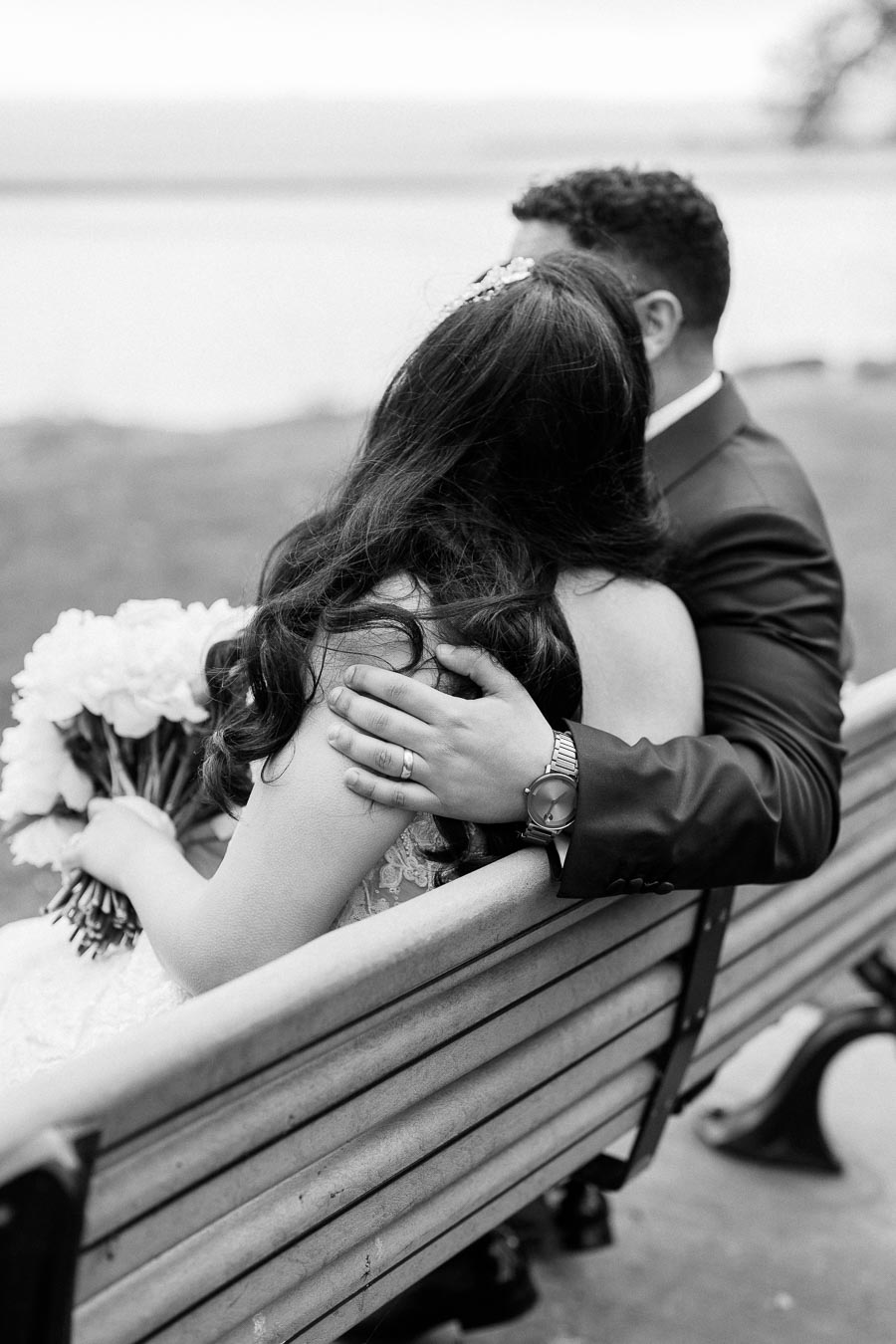 Black and white photo of a newlywed couple sitting on a bench with the bride holding a bouquet, showcasing a romantic embrace by the groom near a serene waterfront.