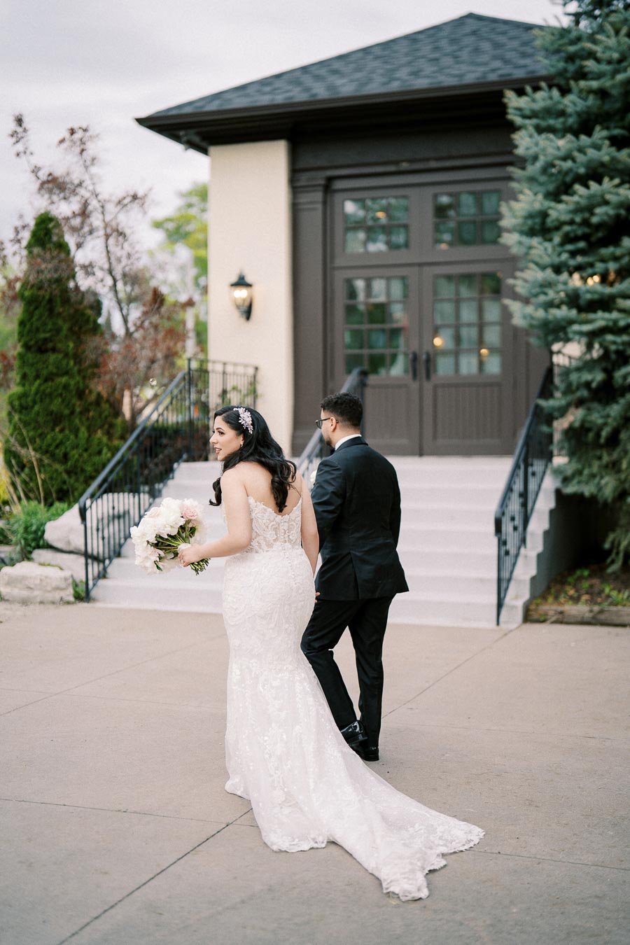 A bride in a white lace wedding dress and a groom in a black suit walking towards a building with a staircase, surrounded by greenery and trees, on a cloudy day.