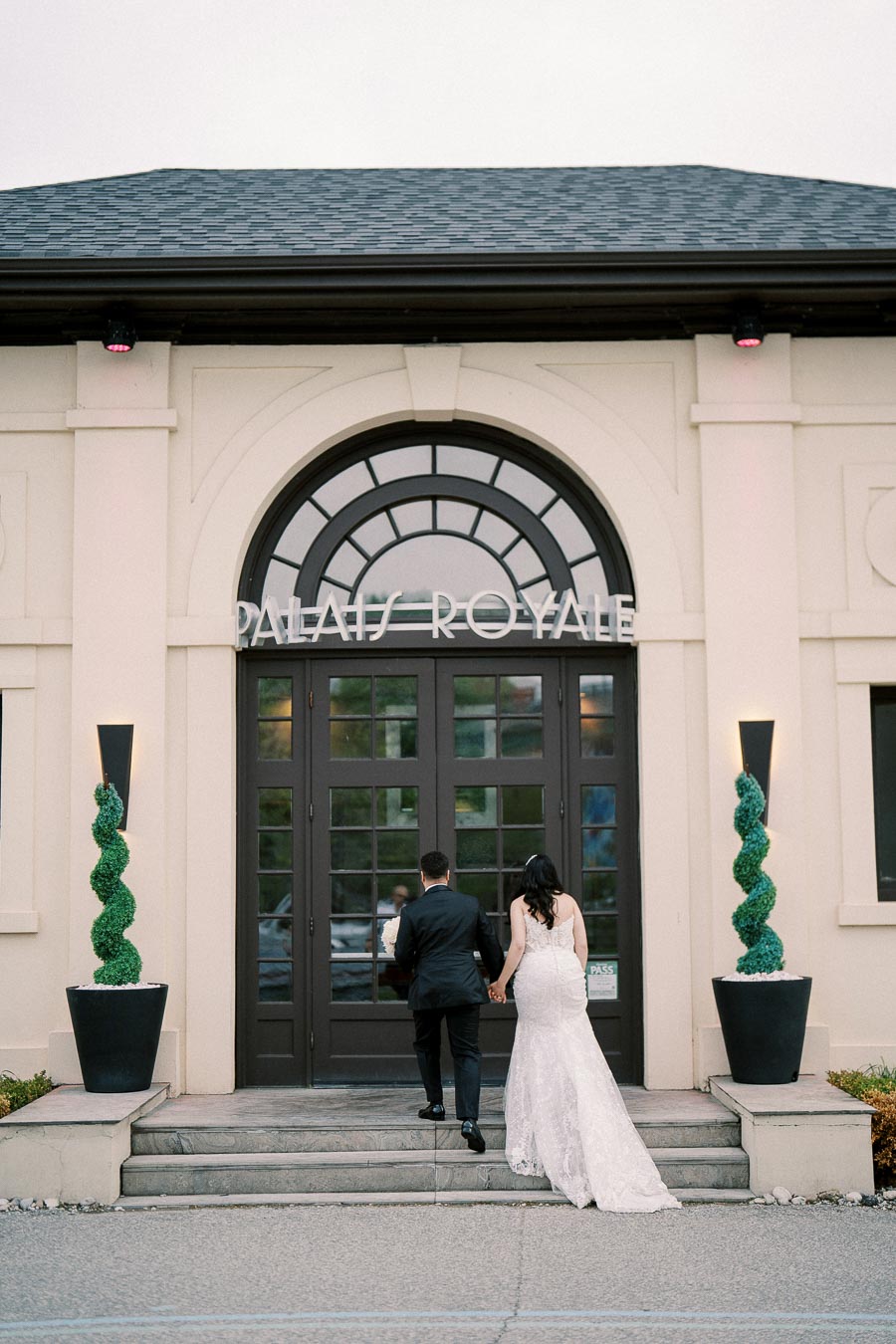 A couple in wedding attire entering Palais Royale event venue with elegant architecture and decorative greenery.