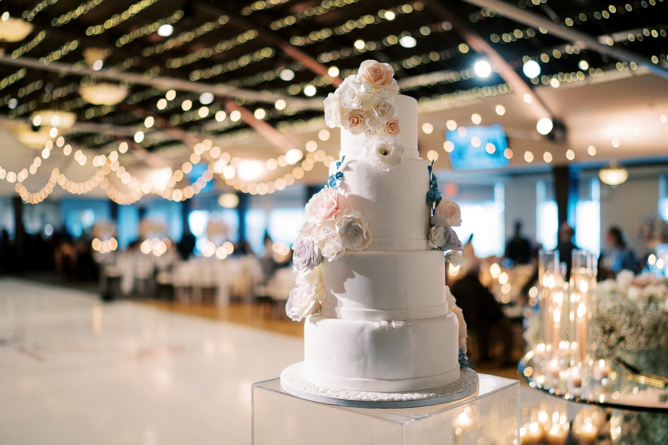 Alt text: Elegant four-tier white wedding cake adorned with pastel flower decorations, set in a beautifully lit reception hall with string lights and softly blurred guests in the background.