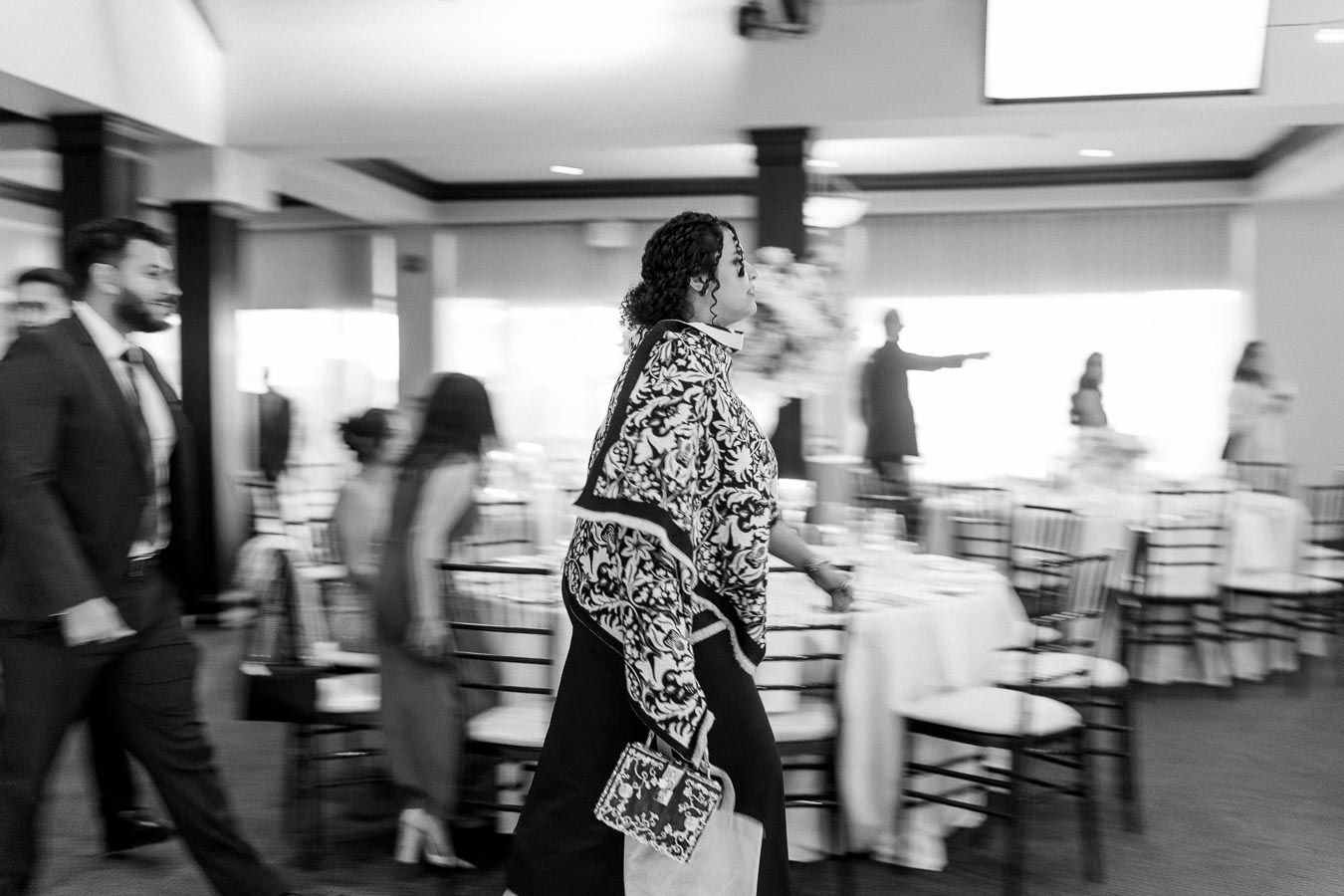 A black and white image of a woman in a patterned blouse holding a decorative handbag walking briskly through a formal dining area, with blurred figures and set tables in the background, conveying a sense of motion and anticipation at a social gathering or event.