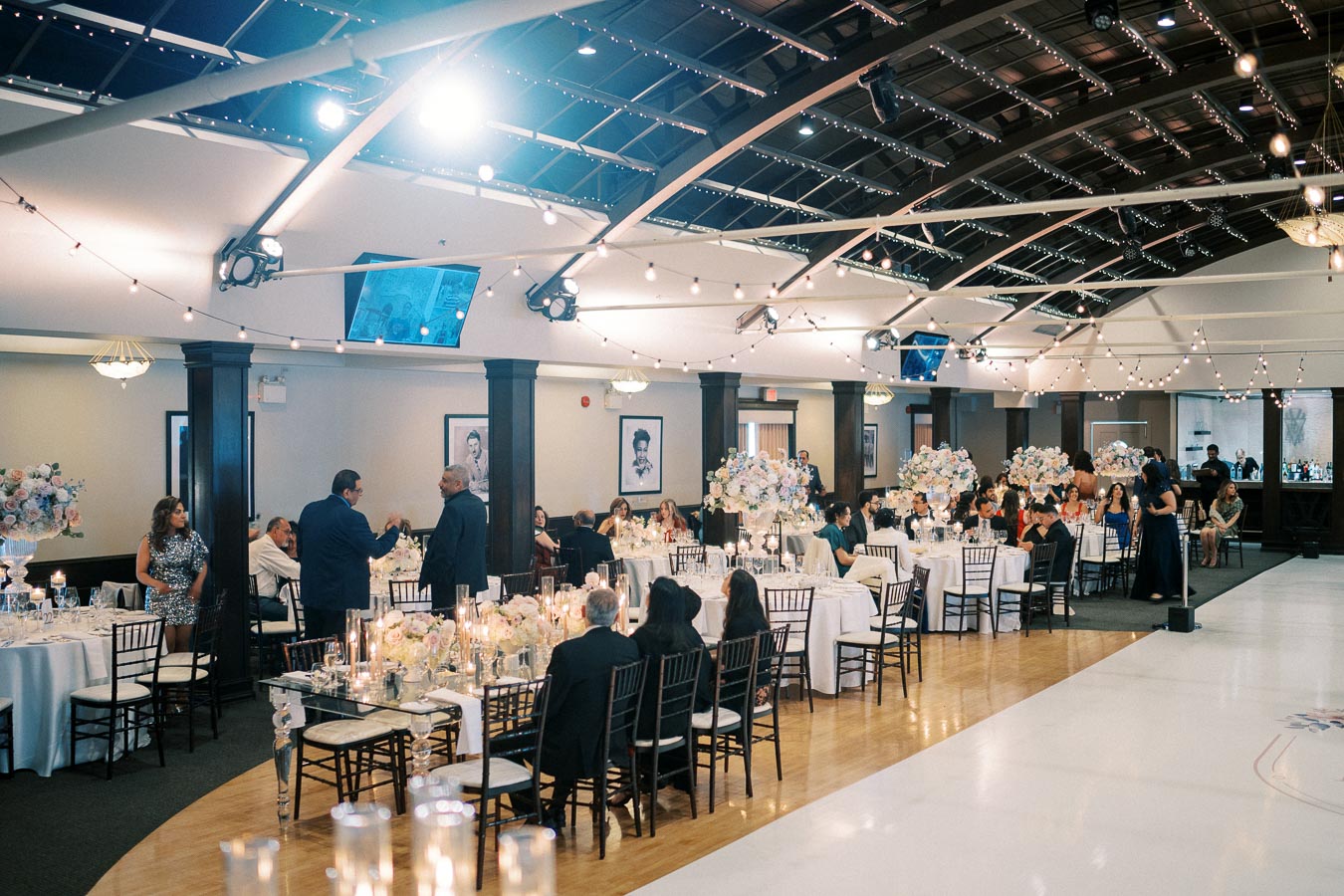 Elegant indoor wedding reception with guests seated at decorated tables under a vaulted ceiling adorned with string lights, creating a festive and sophisticated atmosphere.