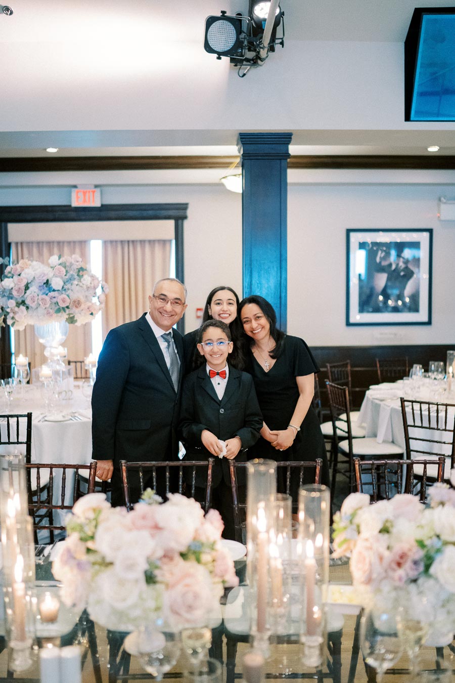 Family posing together at a formal event in an elegantly decorated venue with white and pink floral arrangements and candlelit tables.