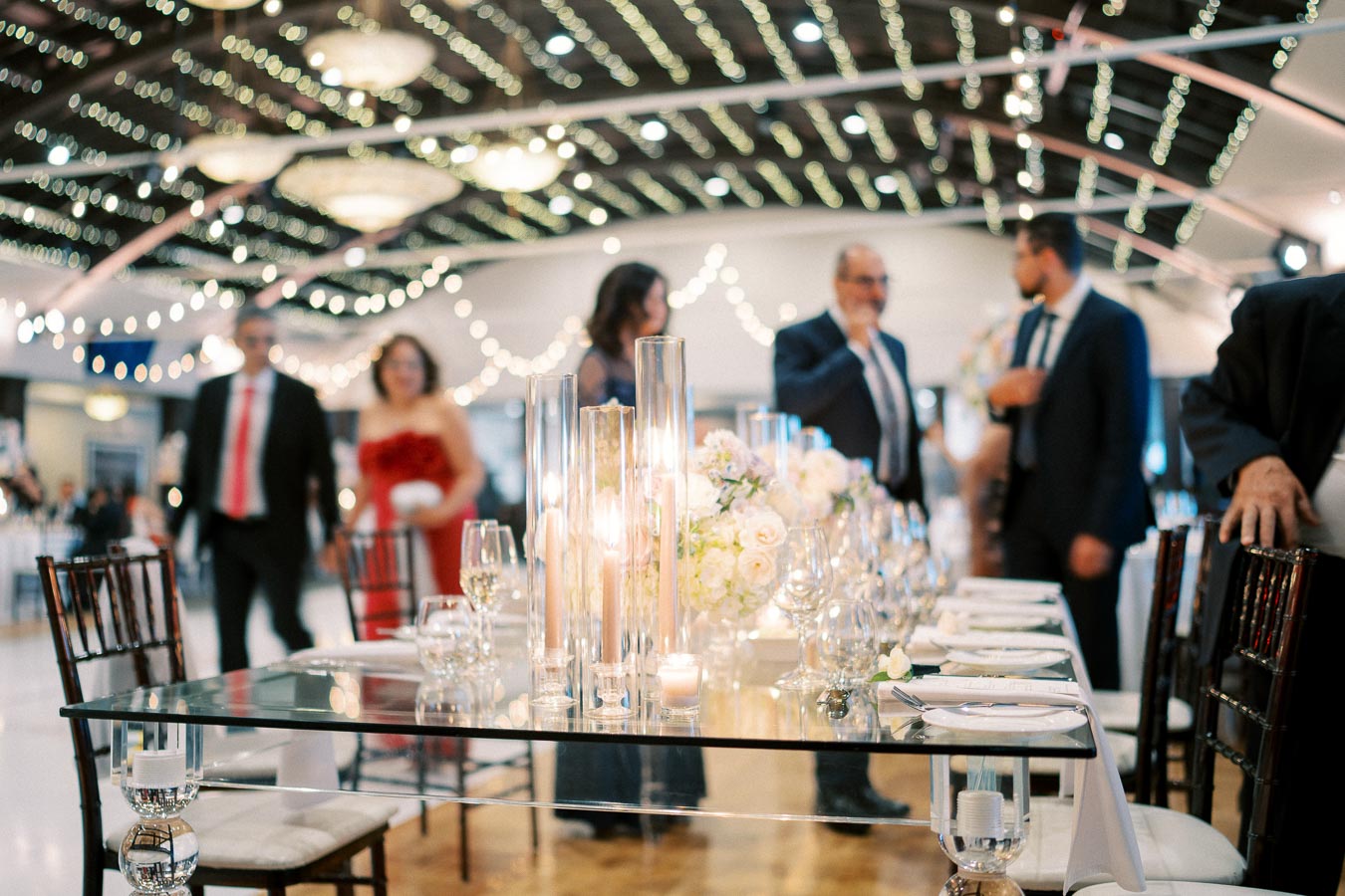 Elegant wedding reception with a beautifully set table featuring glassware, floral centerpieces, and candles, under string lights and a decorative ceiling, with elegantly dressed guests in the background.