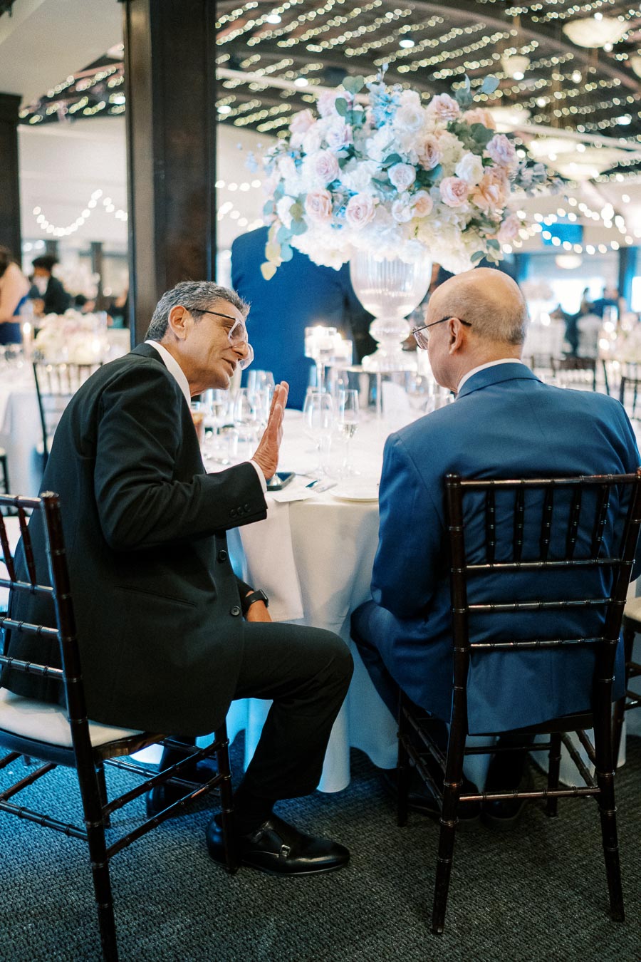 Two men in suits engaged in conversation at an elegantly decorated wedding reception, with a large floral centerpiece and twinkling lights in the background.