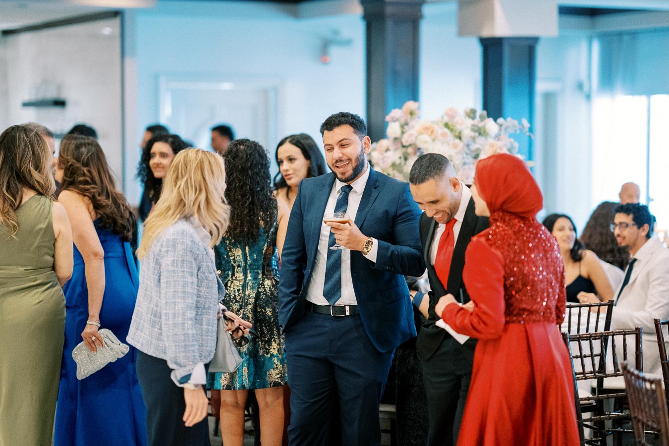 A lively social gathering with diverse people engaged in conversation, elegantly dressed in formal attire, including suits and evening dresses, against a backdrop of floral decorations in a stylish event space.