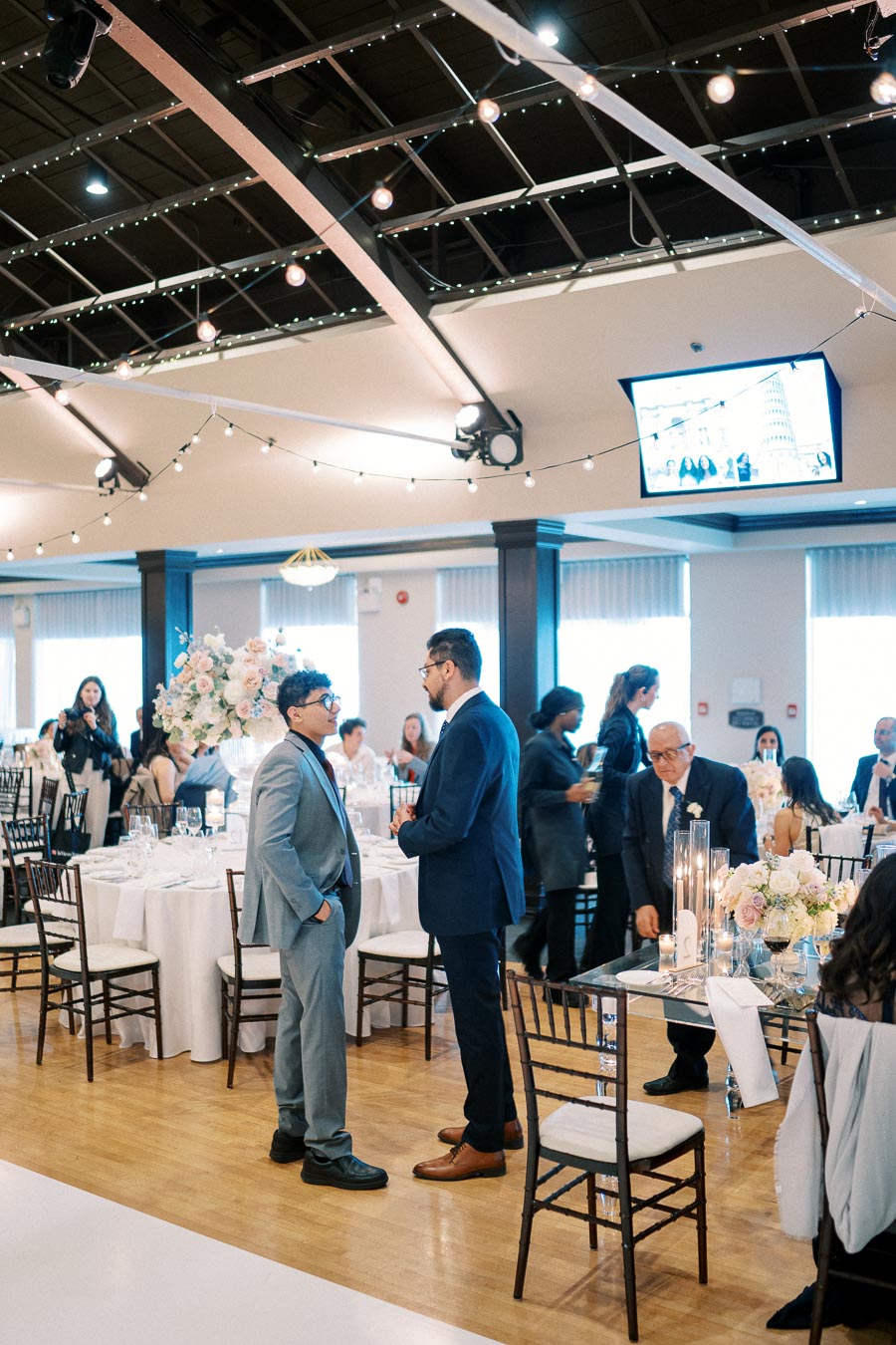 Guests mingling at an elegant indoor wedding reception, with formal attire, beautifully arranged tables, floral centerpieces, and string lights creating a warm atmosphere.