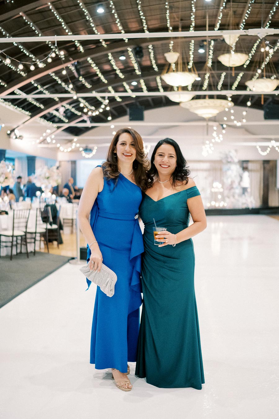 Two women in elegant evening dresses pose smiling at a formal event, with twinkling string lights overhead and a beautifully decorated venue in the background.