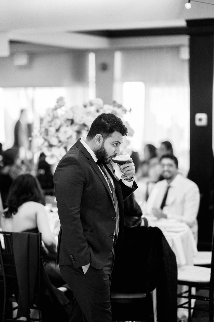 A man in a suit drinks from a glass at an elegant indoor event, with people socializing in the background.