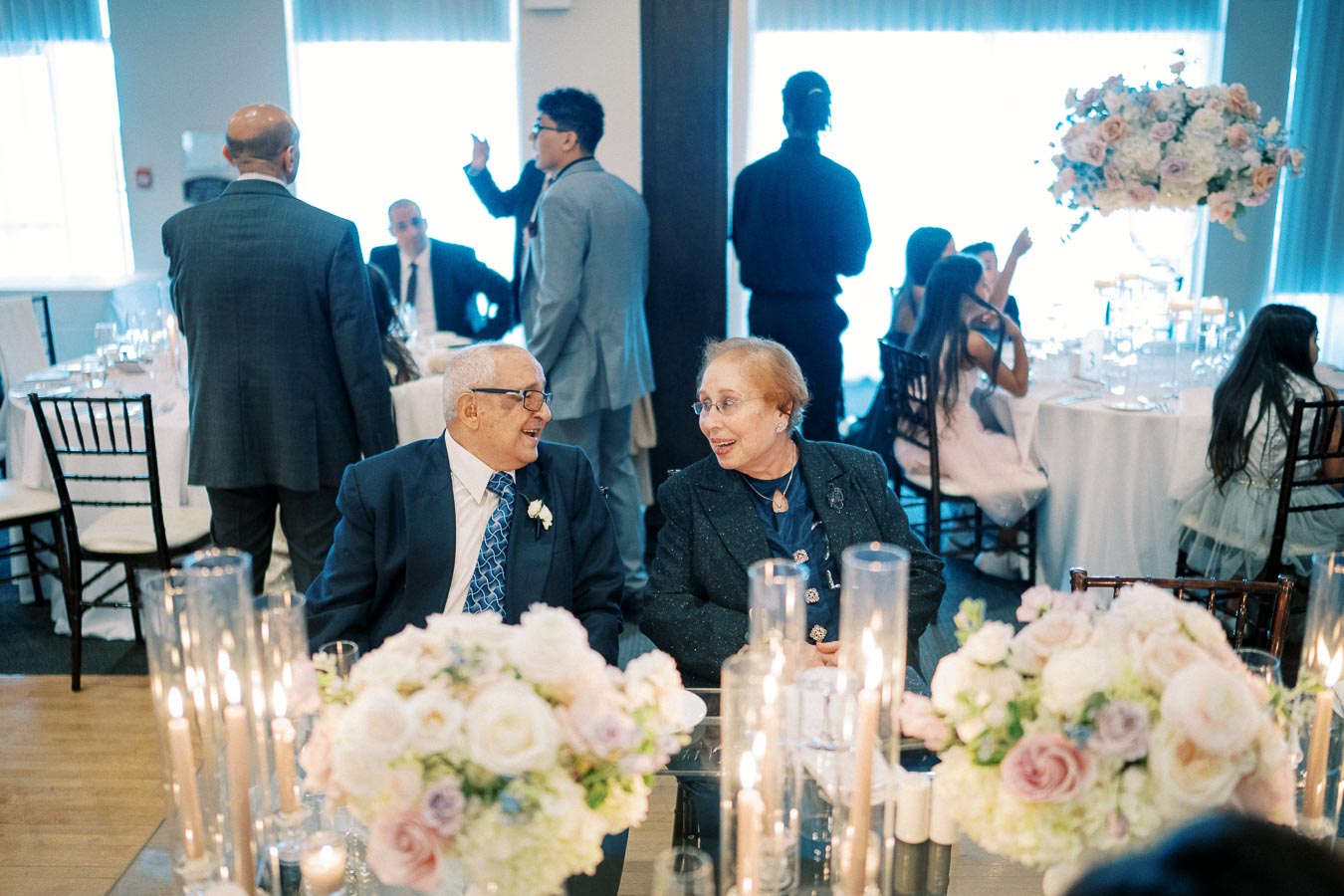 Elderly couple smiling and chatting at an elegant event with floral centerpieces and guests in formal attire.