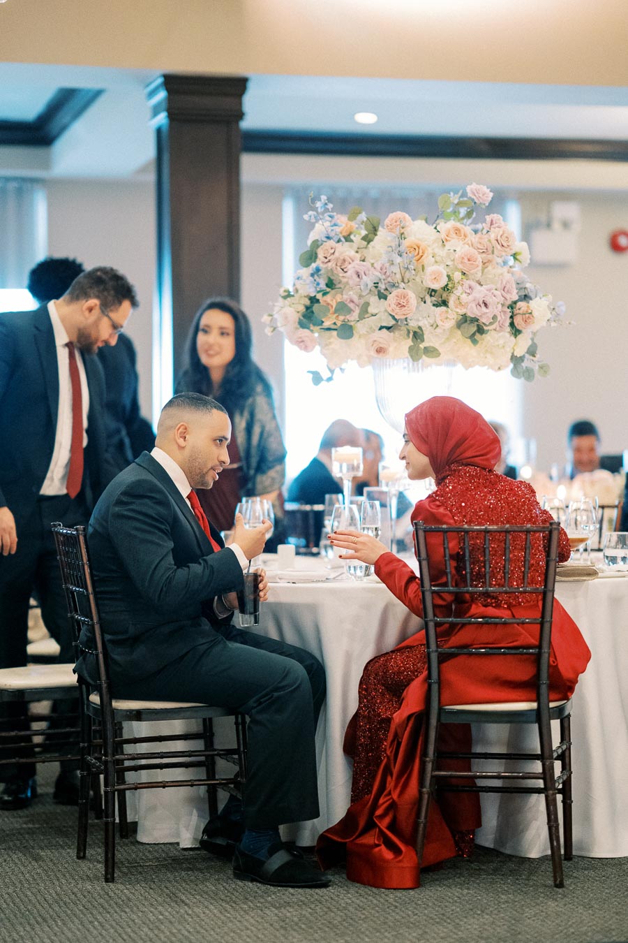 A group of people dressed in formal attire seated at a round table in a banquet setting, with a beautiful floral centerpiece featuring pastel roses and greenery.