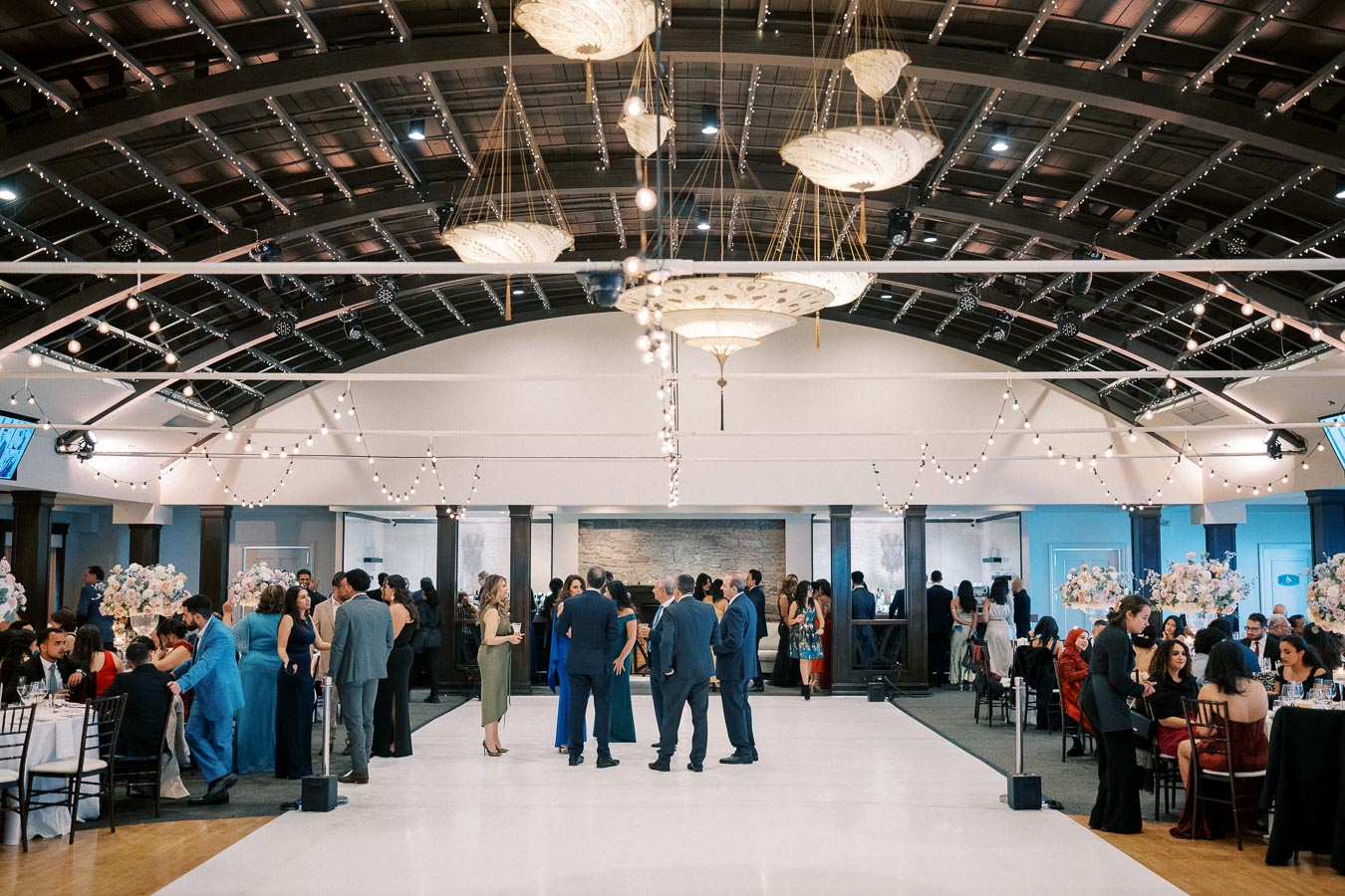 Elegant indoor wedding reception with guests socializing under decorative string lights and ornate chandeliers, featuring beautifully arranged flower centerpieces and a spacious dance floor.