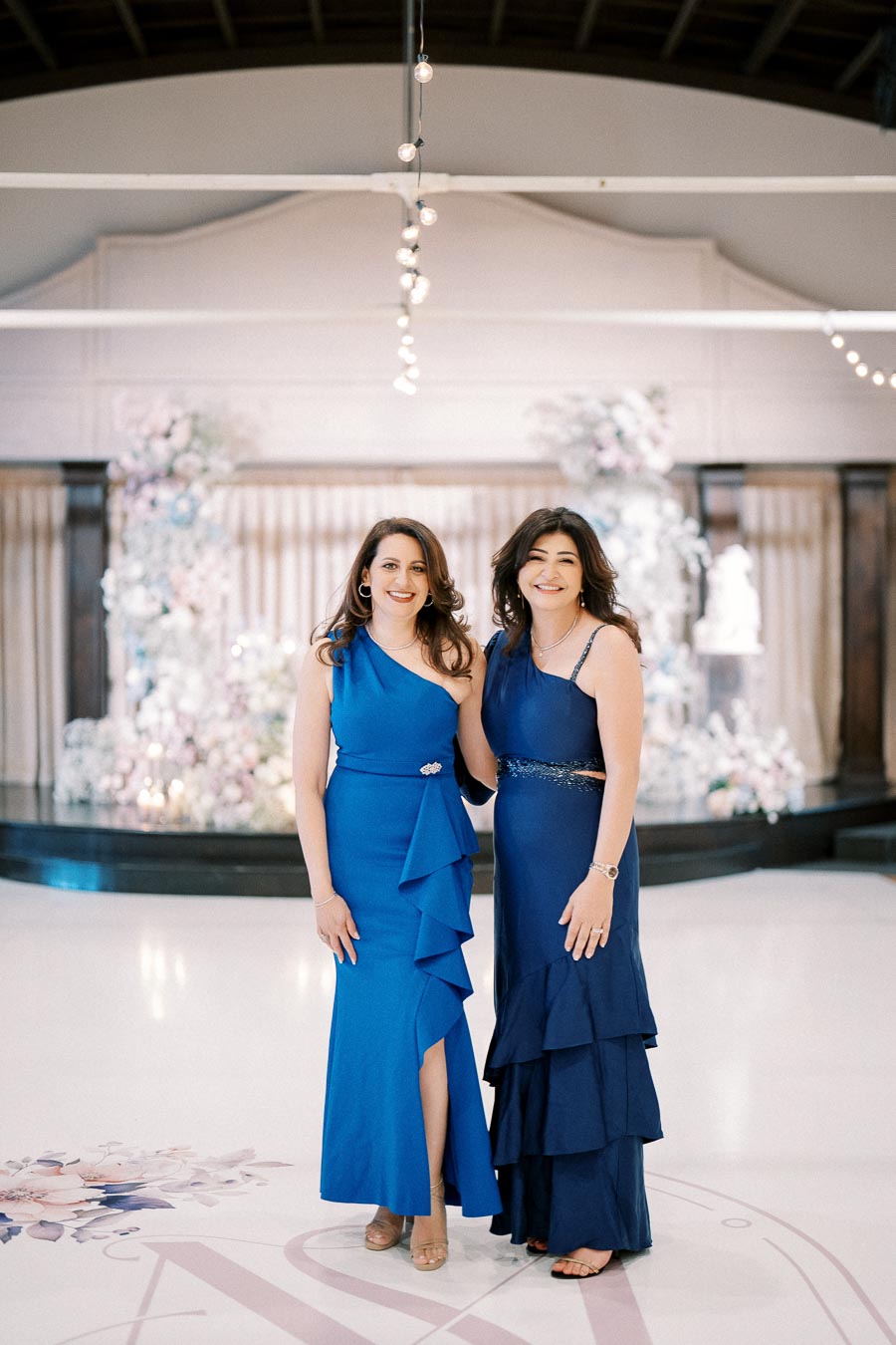Two women in elegant blue dresses pose together in a beautifully decorated indoor venue with floral arrangements in the background.