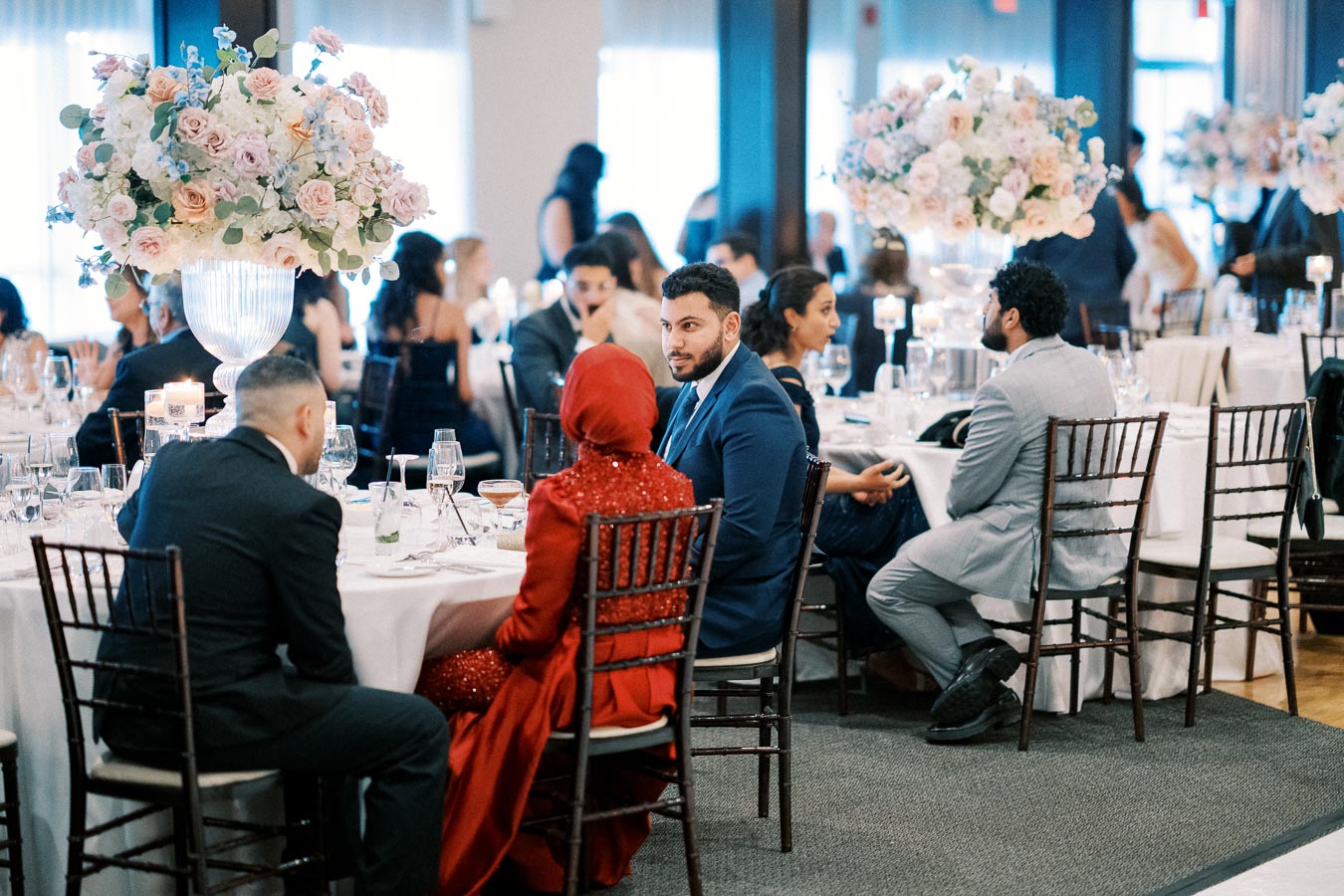 A sophisticated wedding reception with guests seated around elegantly decorated tables adorned with large floral centerpieces, creating a luxurious and festive atmosphere.