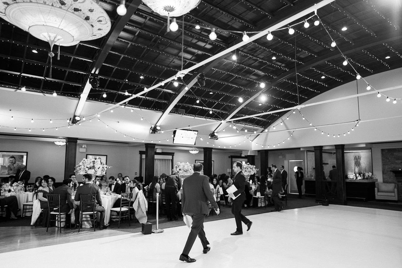 Black and white image of an elegant indoor wedding reception with guests seated at decorated tables, ambient string lights hanging from the ceiling, and two people walking across the dance floor.