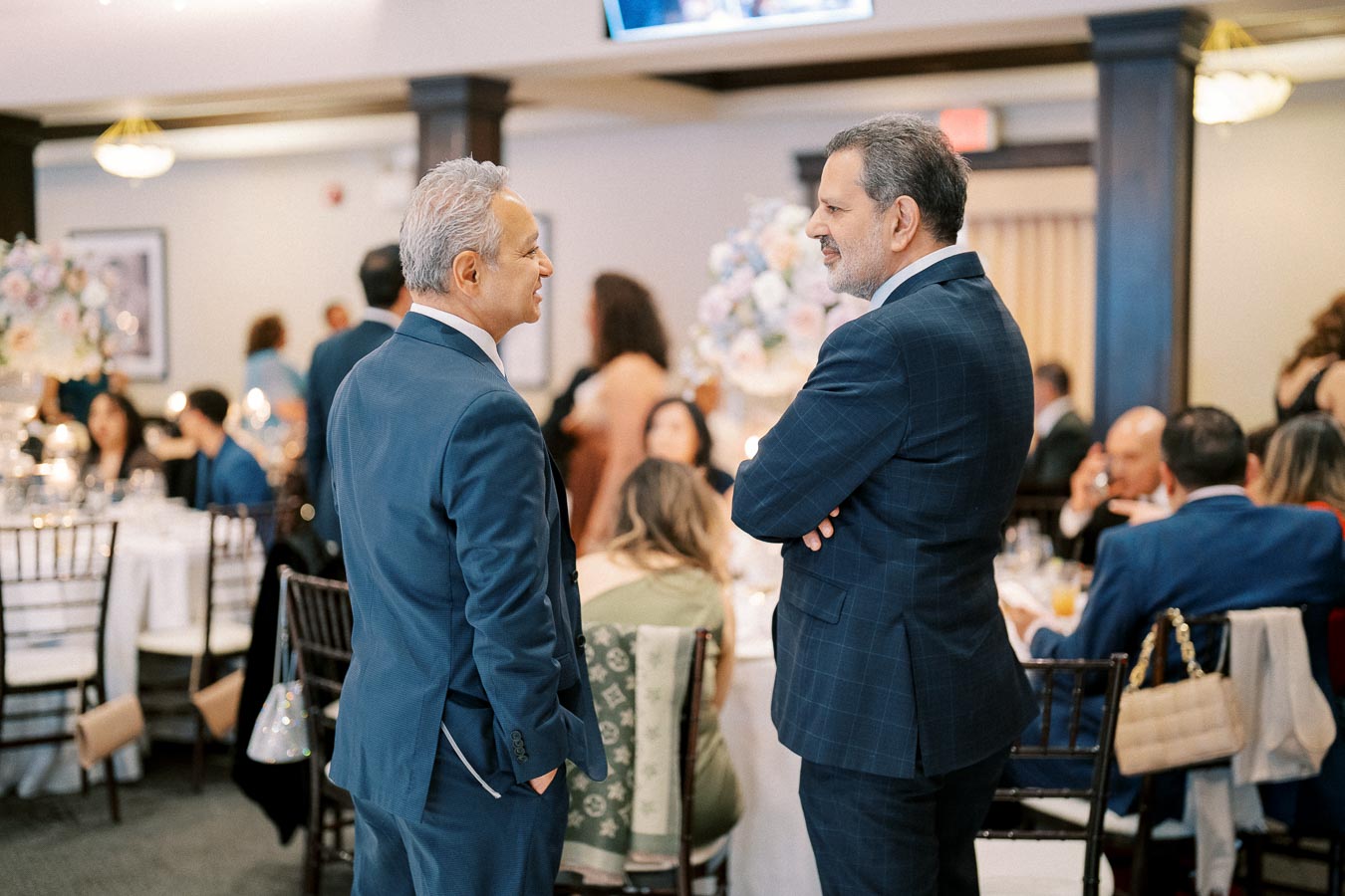Two men in suits conversing at a formal event, with a seated audience and elegant table settings in the background.