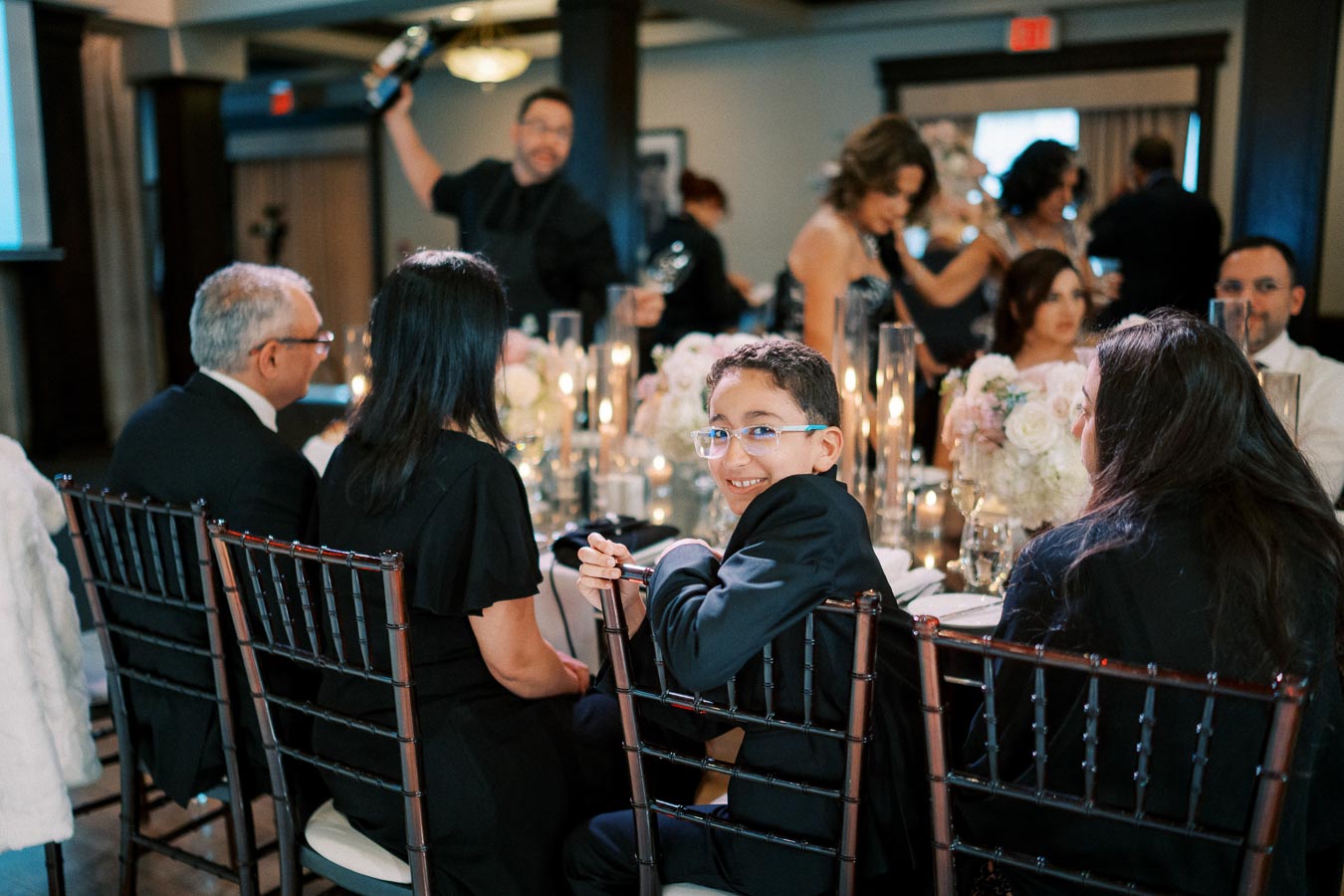 A joyful wedding reception scene featuring a smiling young boy in glasses seated at a beautifully decorated table with flowers and candles, surrounded by elegantly dressed guests in a warmly lit venue.